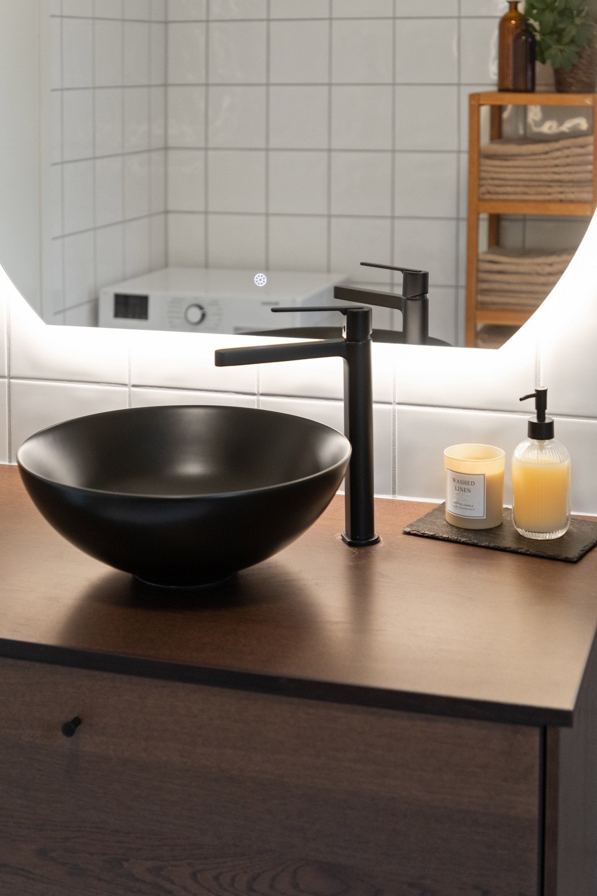 Et stilig speil med bakenforliggende belysning er plassert over en stor benk med mørke skuffer og toppmontert servantskål i sort design Galleribilde