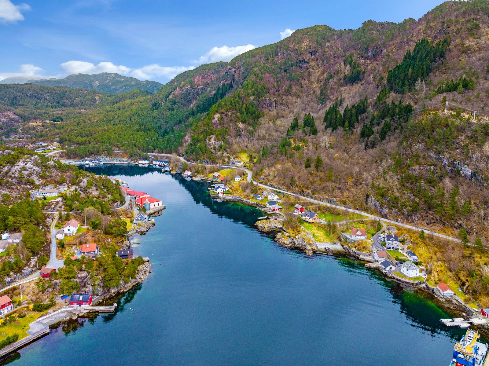 Liten fjord med hus langs sjøkanten, omgitt av skogkledde åser og fjell. Galleribilde