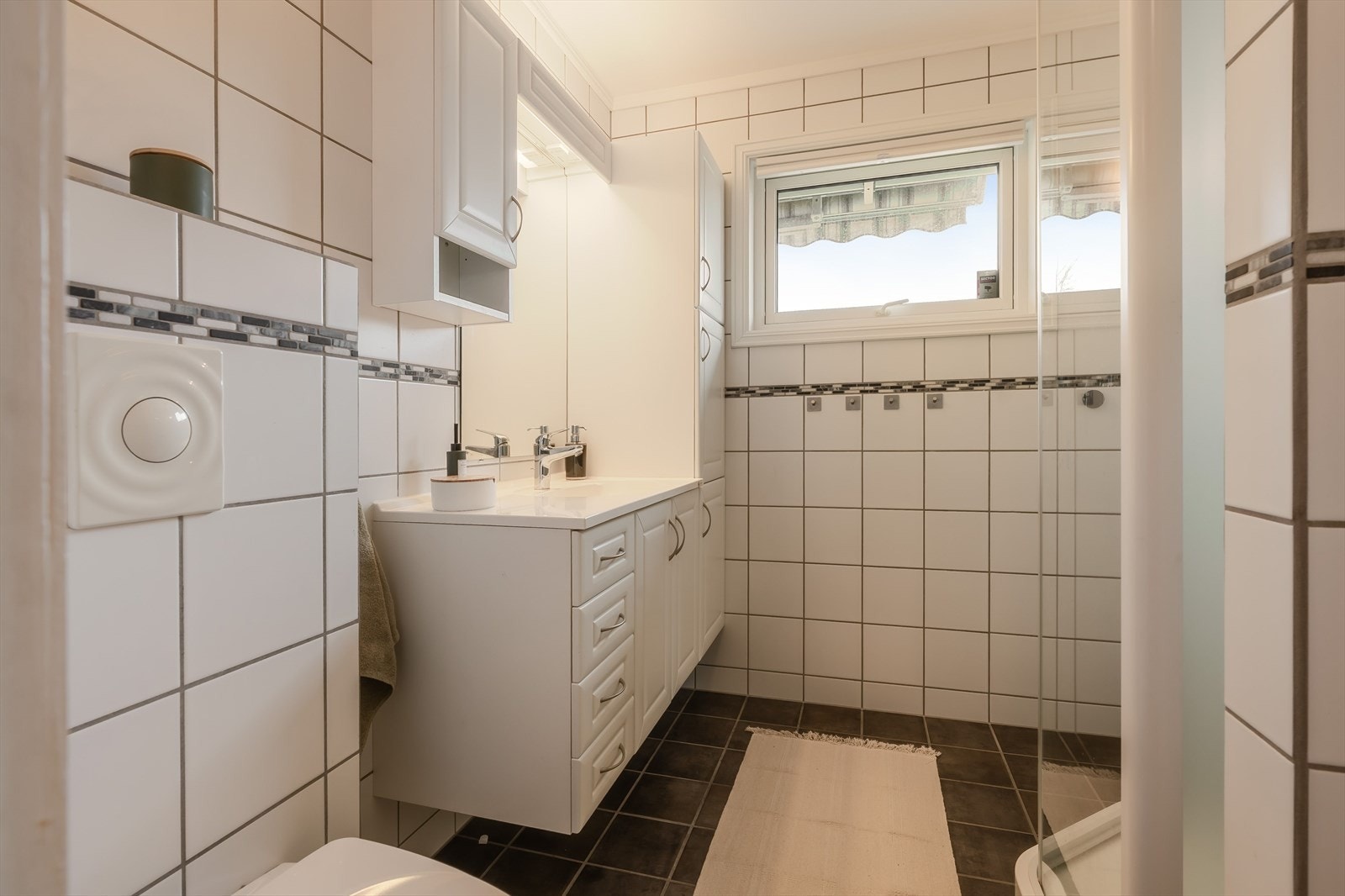 Badet i denne etasjen er flislagt med hvite fliser på vegger og sorte fliser på gulv. Her er det innredet med dusjkabinett, servant med tilhørende oppbevaringsløsning og toalett. Galleribilde