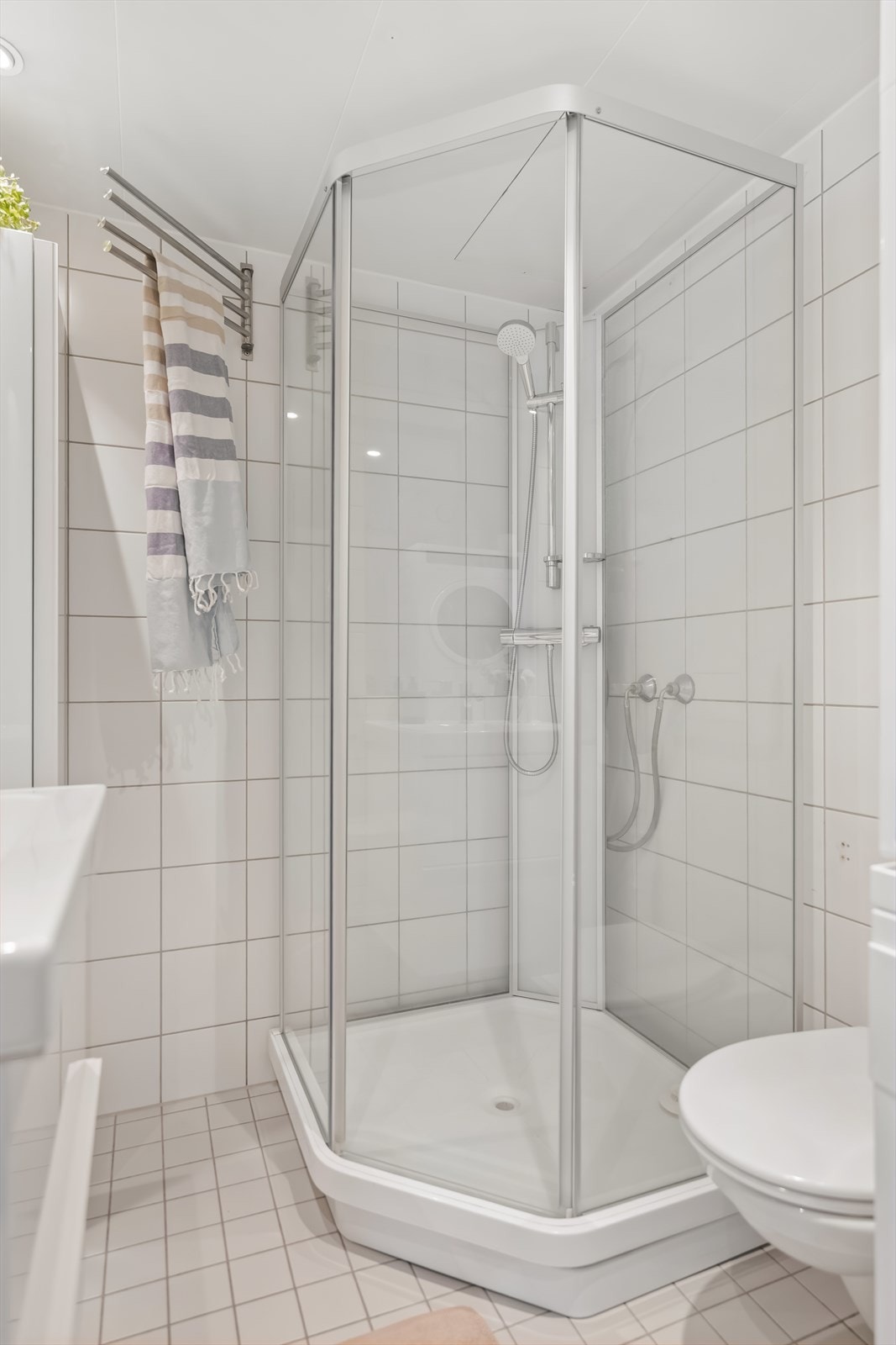 Badet har glatte høyglansfronter og heldekkende servant. Det er innmurt speil over servanten, veggmontert klosett, opplegg for vaskemaskin samt dusjkabinett med skyvedør og dusjarmatur. Galleribilde