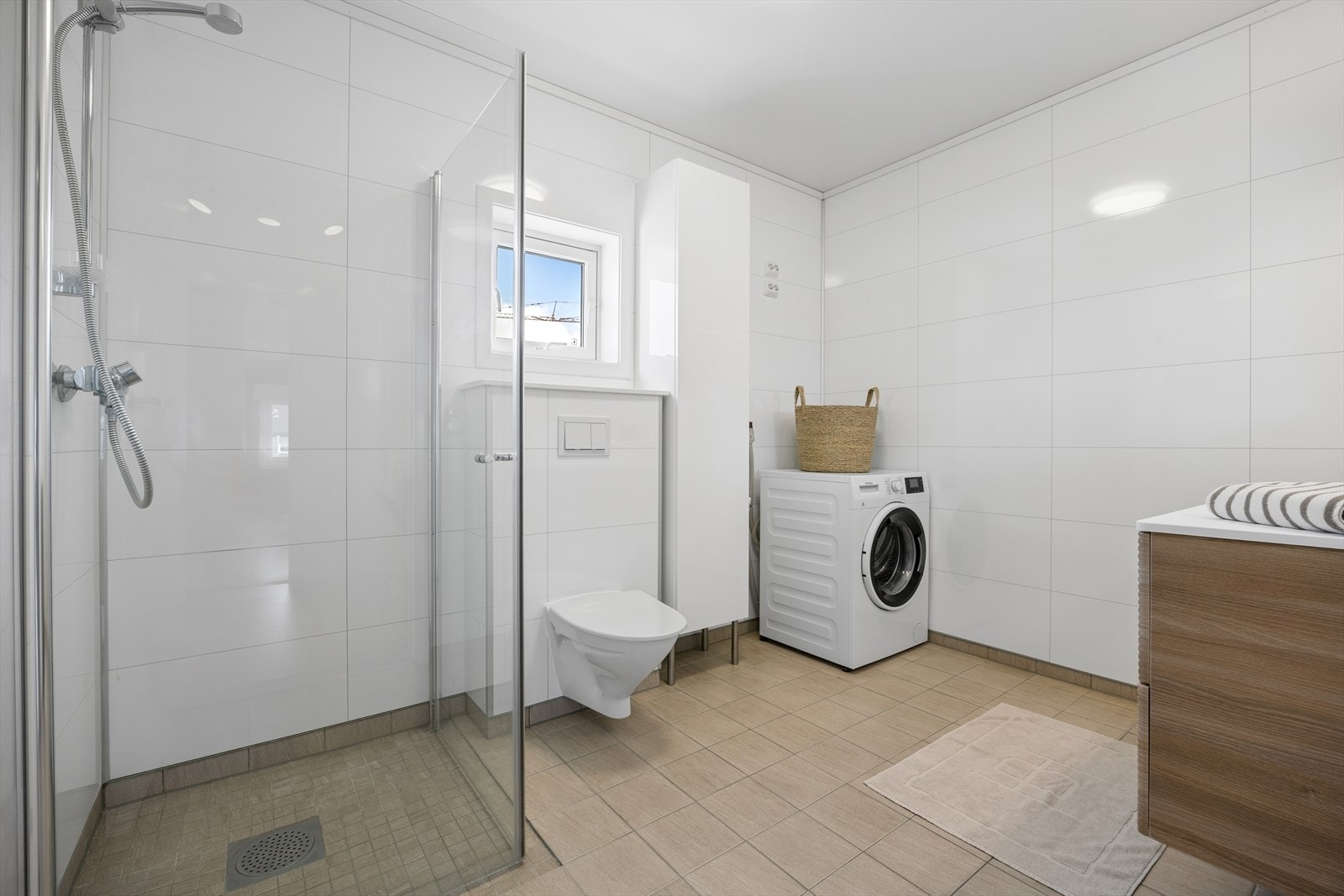 Badet har flislagt gulv, dusj-sone, veggmontert toalett, opplegg for vaskemaskin/tørketrommel og en stilig baderomsinnredning. Galleribilde