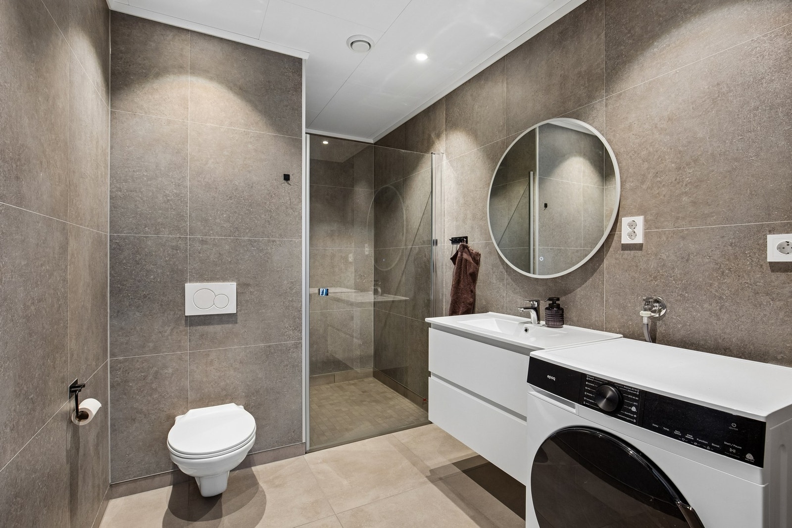 Badet har innredning med nedfelt servant, veggmontert toalett, dusjkabinett og opplegg for vaskemaskin Galleribilde