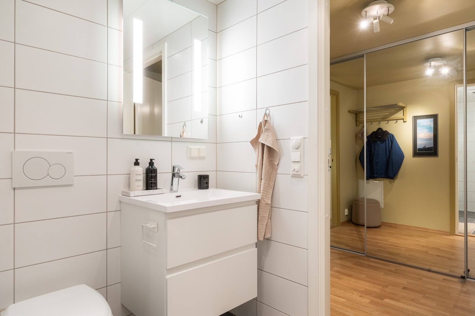 Videre er badet innredet med vegghengt toalett, servant med innredning, speil med belysning og dusjhjørne med svingbare glassdører. Galleribilde