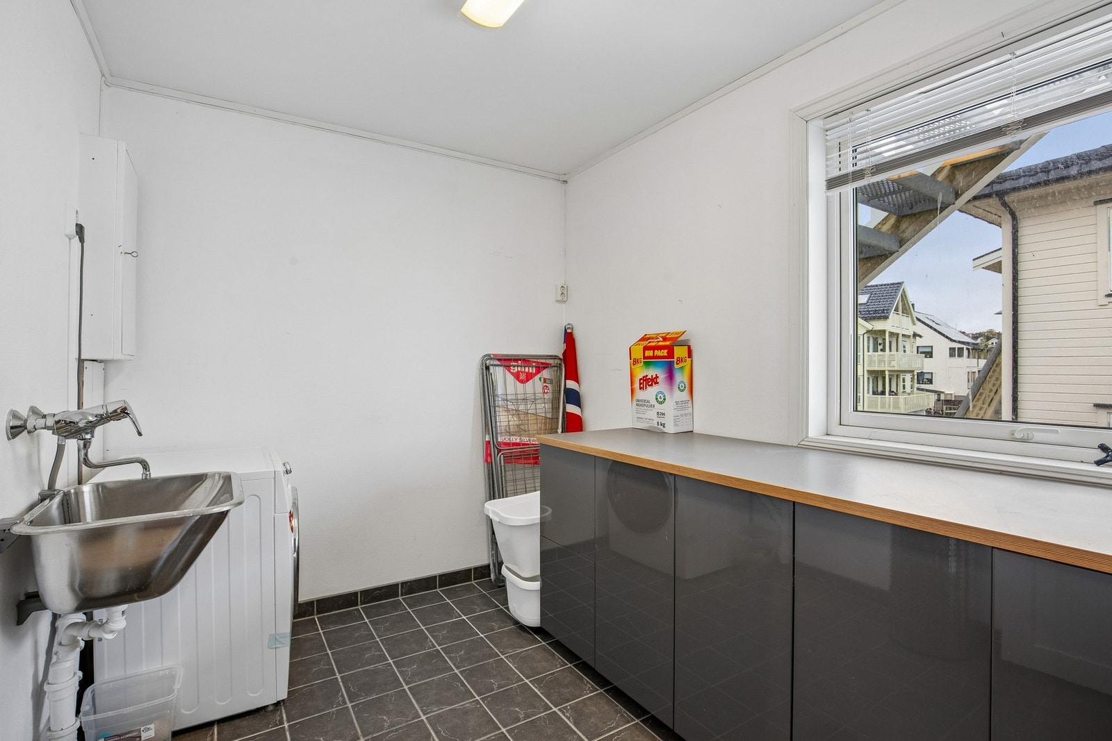 Praktisk vaskerom med flislagt gulv, opplegg for vaskemaskin samt utslagsvask. Det er satt inn benkeskap som gir god lagrings- og arbeidsplass. Galleribilde