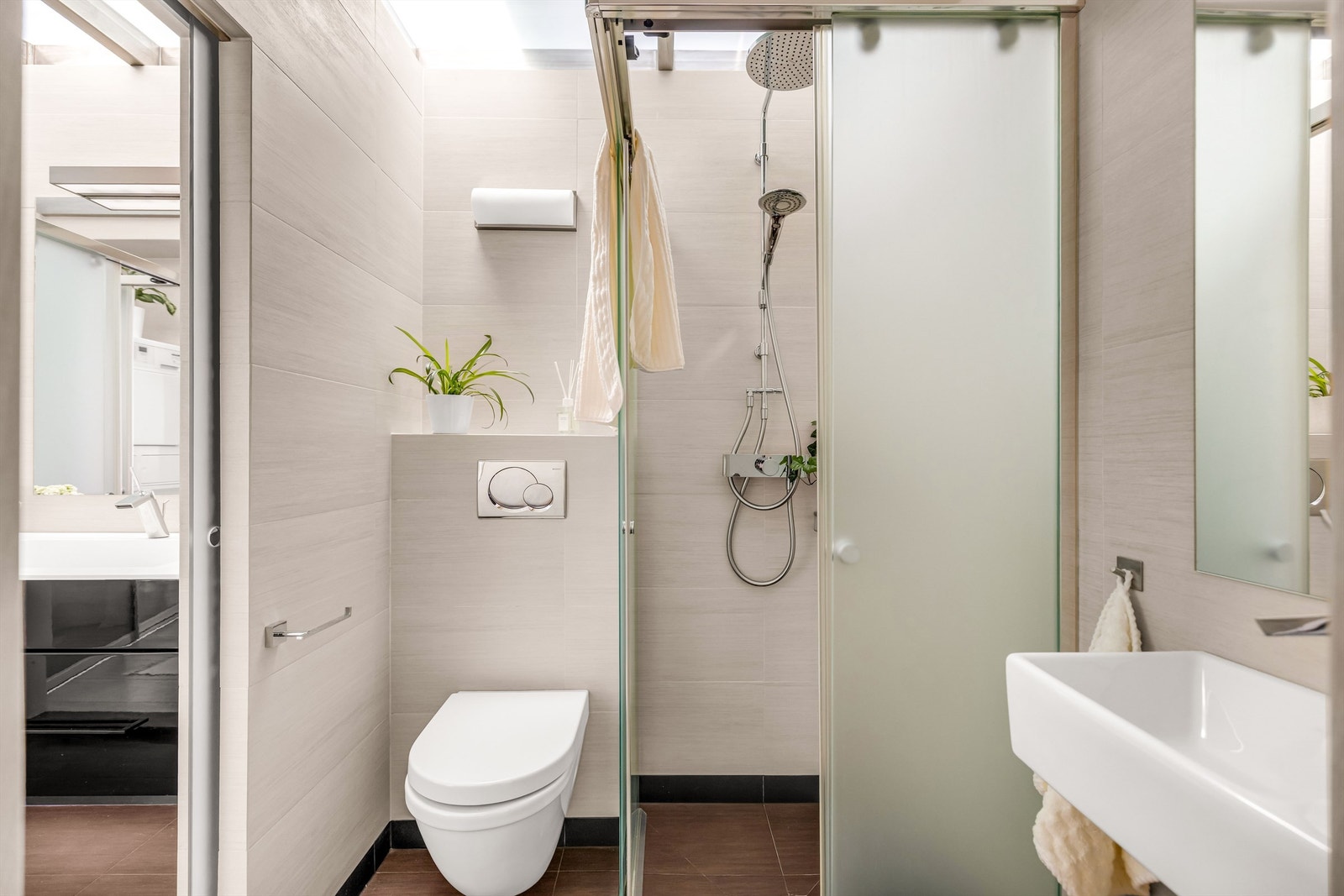 Badet nærmest gangen er innredet med vegghengt toalett og dusjnisje i frostet glass, to-greps dusjarmatur på vegg og regnfallsdusj Galleribilde