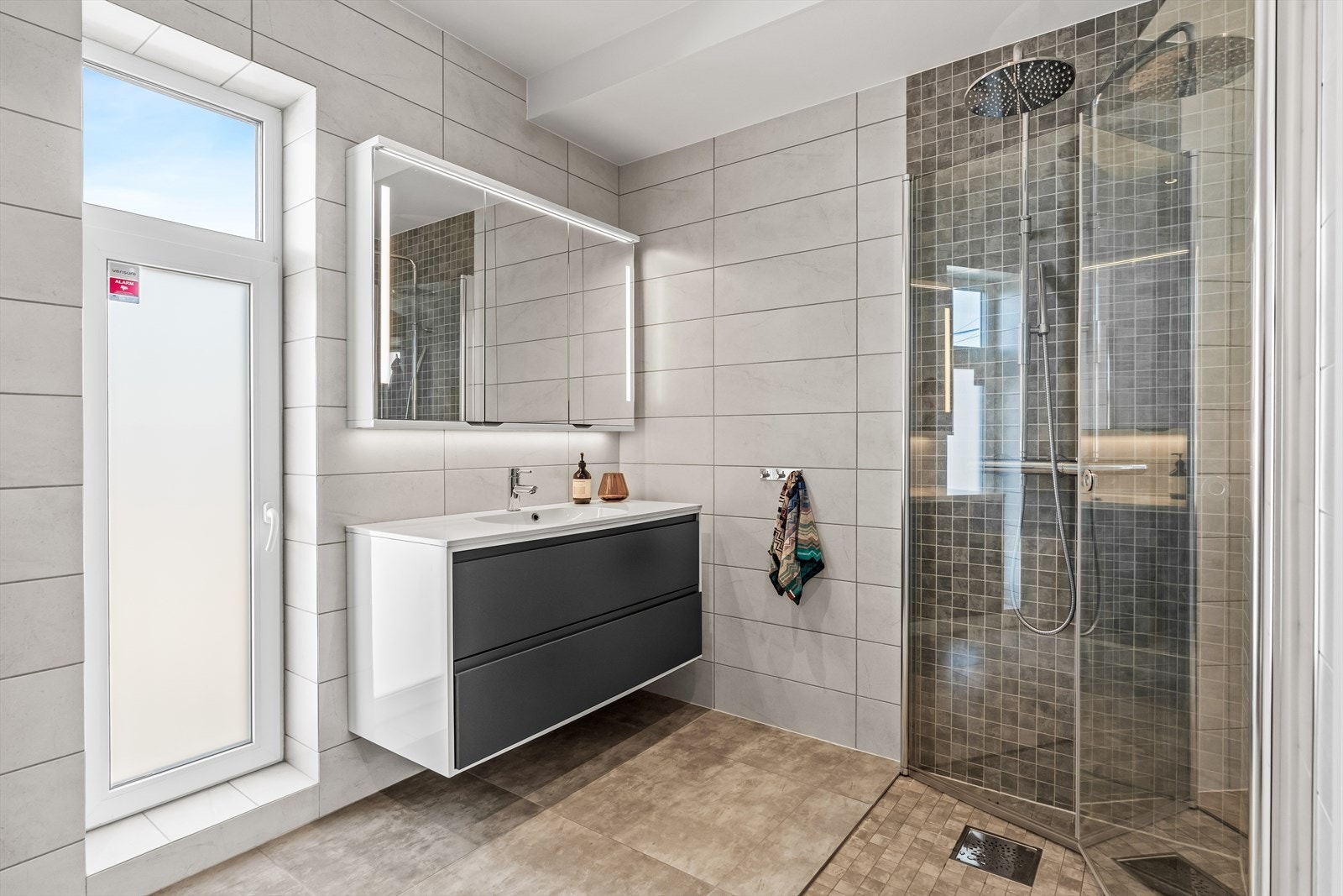 Badet i første etasje har gulv med store fliser og mosaikk i grånyanser, lyse veggfliser og gulvvarme. Galleribilde