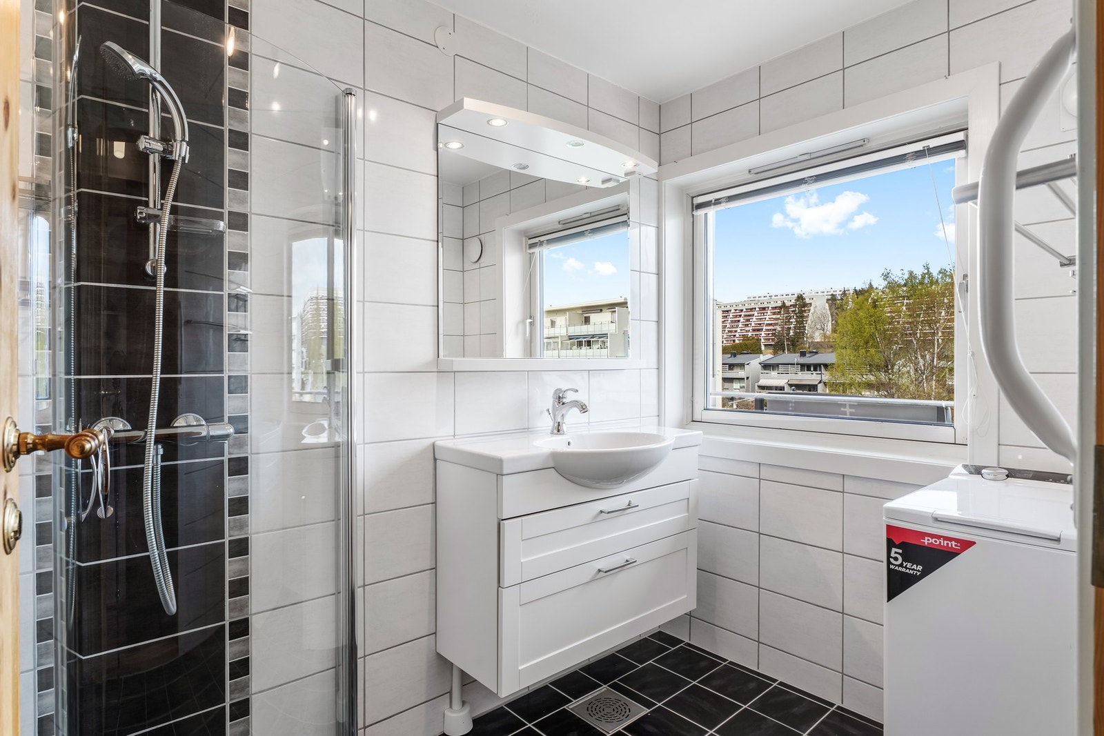 Lyst bad med flislagte overflater, speil over servant og opplegg for vaskemaskin, samt mekanisk avtrekk. Galleribilde