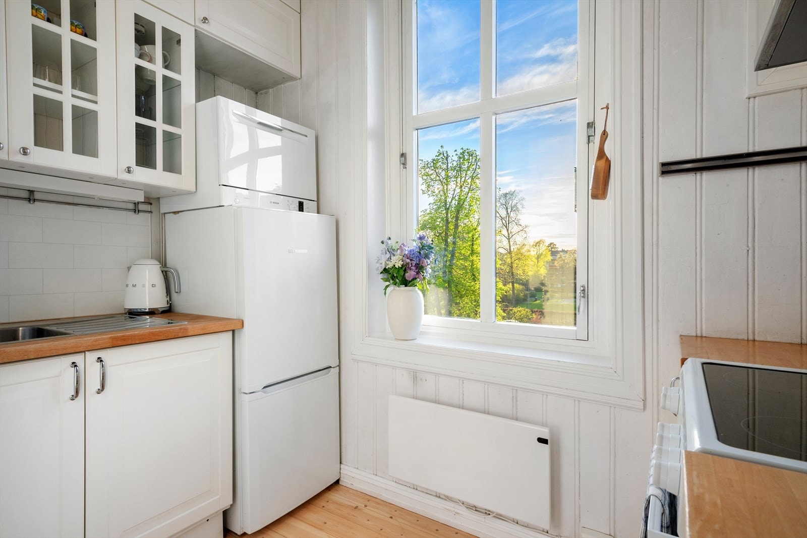 Kjøkkeninnredning med profilerte fronter (fornyet i 2023) og glassfelt i overskap, som gir et klassisk uttrykk. Heltre benkeplate og nedfelt kum i rustfritt stål. Galleribilde