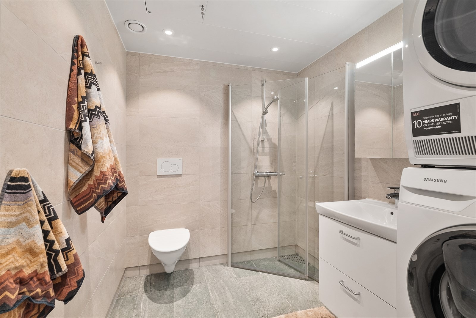 Delikat flislagt bad med vannbåren gulvvarme. Badet er utstyrt med dusjhjørne, vegghengt toalett og opplegg for vaskemaskin/tørketrommel. Galleribilde