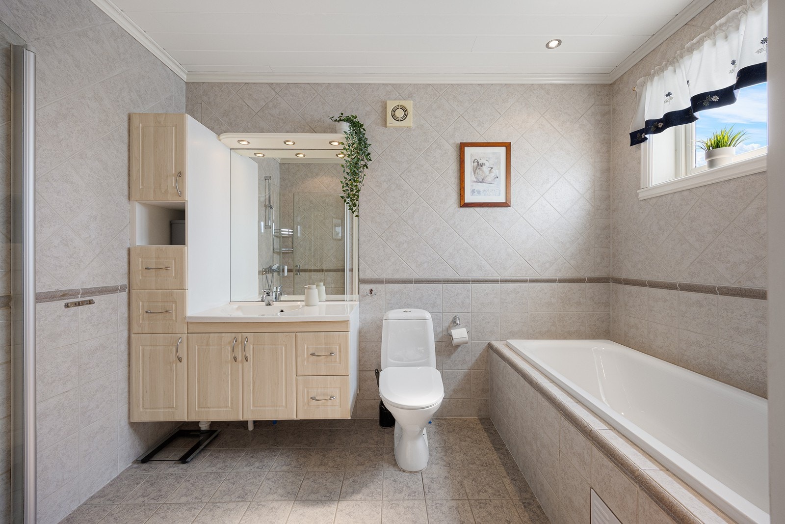 Baderomsinnredningen består av et lyst servantskap, et matchende høyskap og et speil med belysning over servanten. Varme er lagt i gulvet, og badet har også gulvmontert toalett og downlights i taket. Galleribilde
