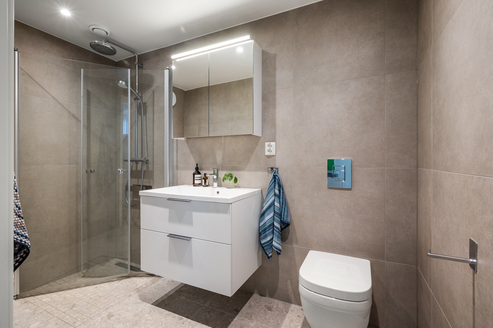 Delikat en-suite bad med store 60×60 cm fliser, vegghengt toalett og servant med speilskap. Galleribilde