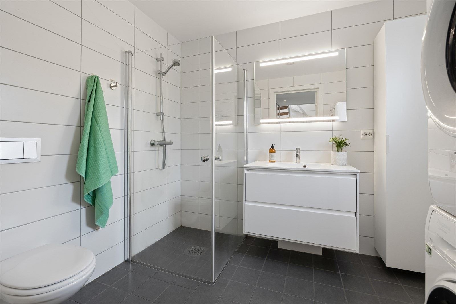 Pent flislagt baderom som har varmekabler i gulvet for økt komfort. Badet er utstyrt med vegghengt toalett, dusjhjørne med glassdører, baderomsinnredning med servant og ett-greps blandebatteri, veggskap, samt opplegg for vaskemaskin. Galleribilde