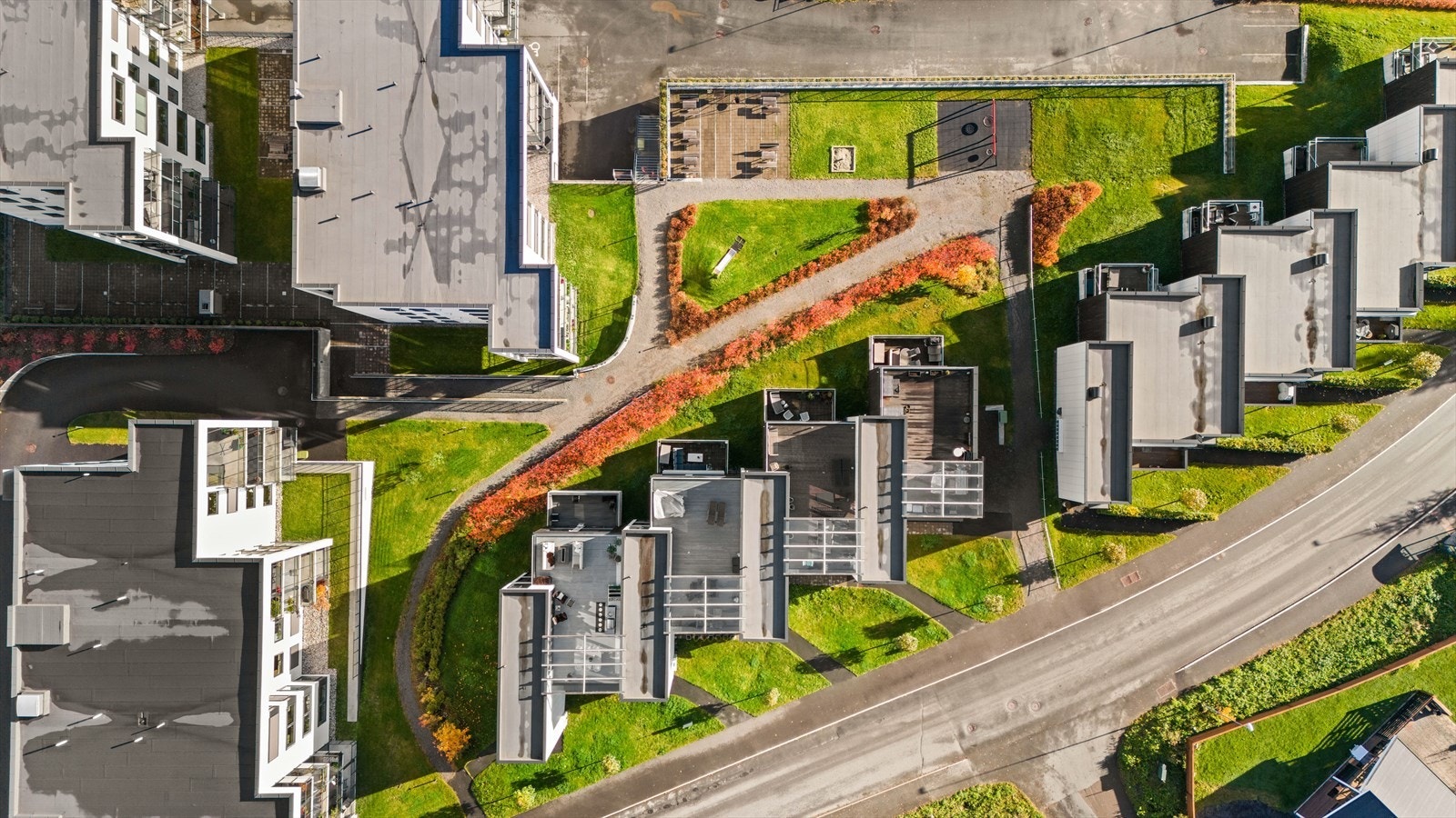Fellestomten er skrånende og opparbeidet med plen, beplantning, sittebenker, lekeapparater og asfaltert gårdsplass. Galleribilde