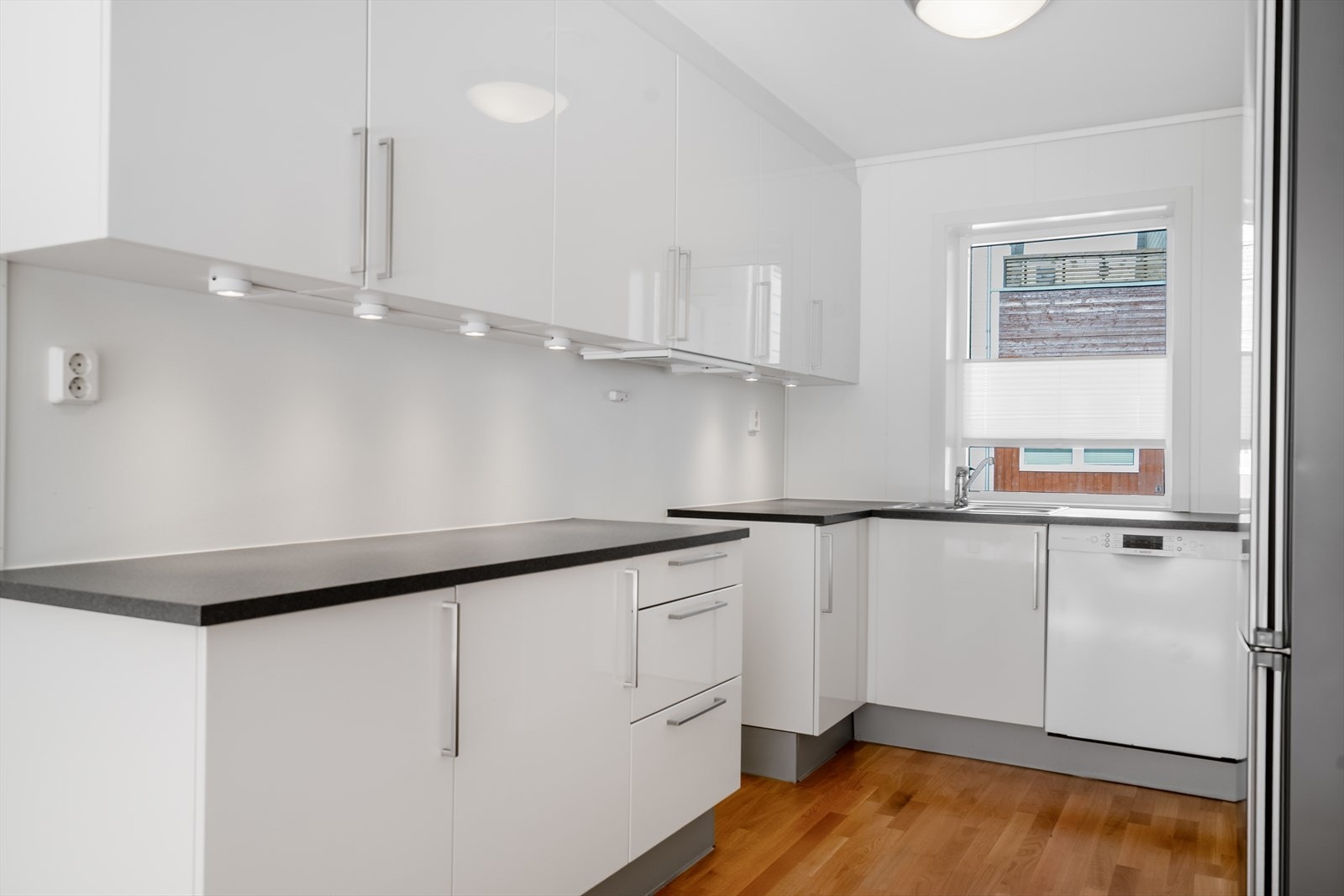 Vinklet kjøkkeninnredning med takhøye overskap. Mørk laminat benkeplate med nedfelt oppvaskbenk og opplegg for oppvaskmaskin. Galleribilde