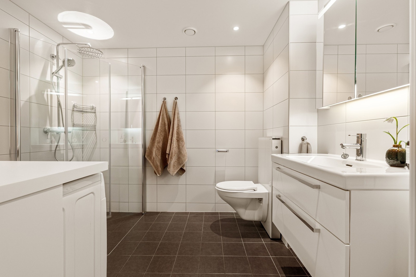 Badet inneholder heldekkende servant med skuffer, speilskap med integrert belysning, veggmontert høyskap, dusjhjørne med innfellbare dører i herdet glass og vegghengt klosett. Galleribilde