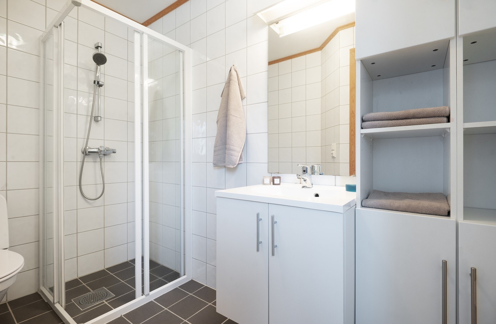 Rommet er utstyrt med gulvstående toalett, dusjhjørne med glass-skyvedører og ettgreps dusjarmatur, servantskap med hvite, glatte fronter og en servant i komposittmateriale. Galleribilde