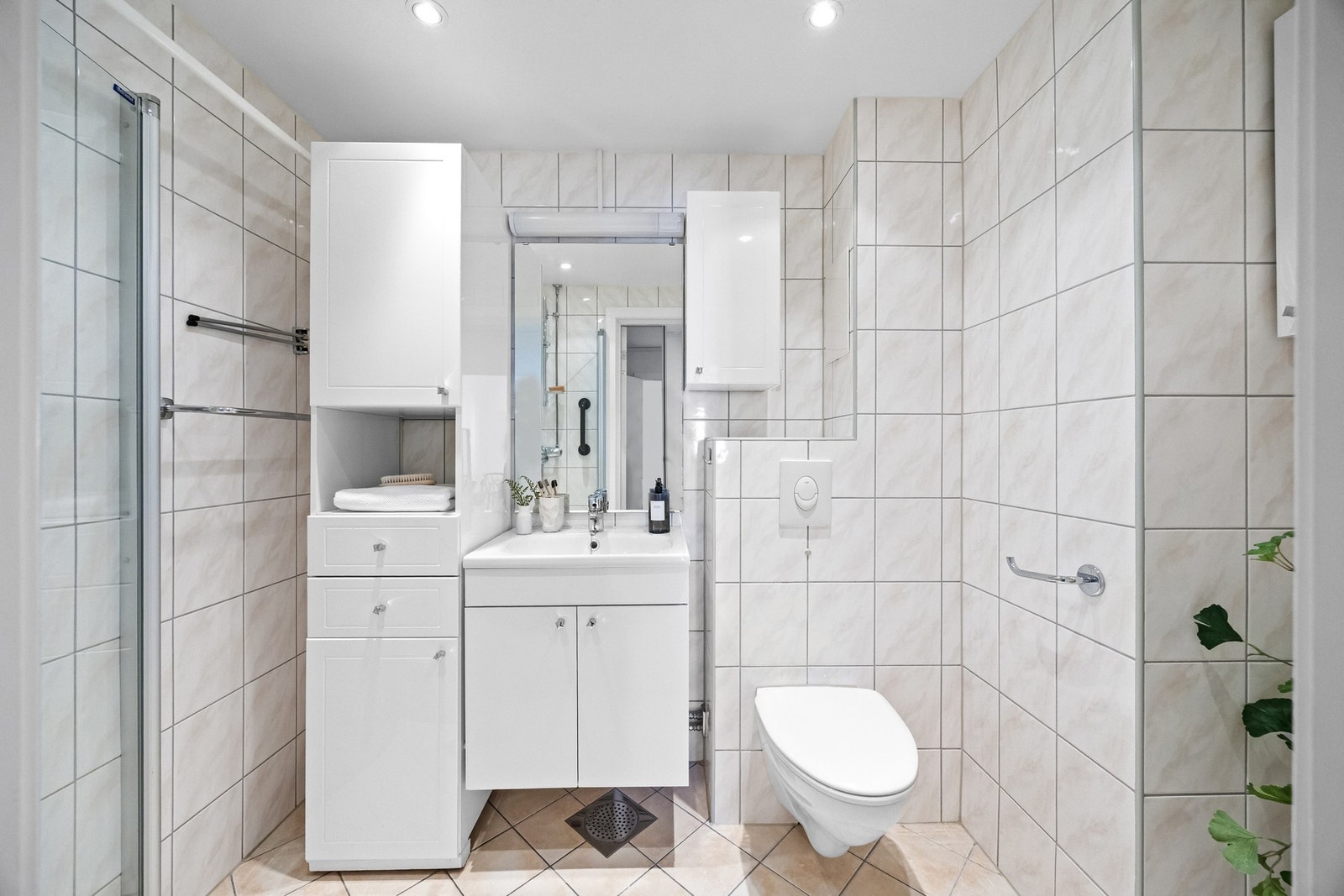 Flislagt bad med varmekabler og downlights. Innredningen består av servantskap, heldekkende servant og speil på vegg. Det er veggmontert klosett og dusjhjørne med svingdør i glass. Galleribilde