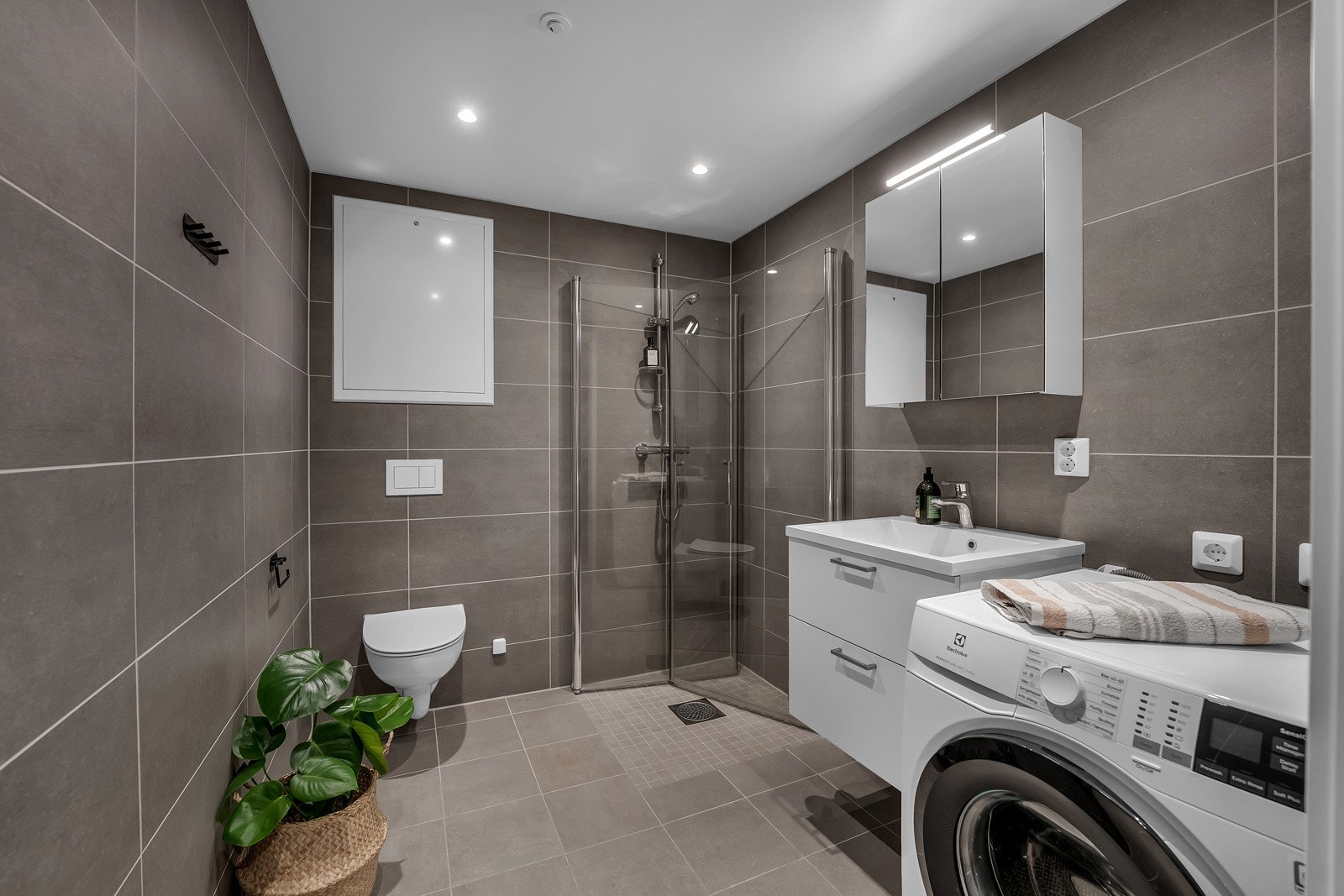 /wc/vaskerom i tilknytting til entreen - med mørkegrå fliser på vegg, downlights i himlingen og lysegrå fliser på gulv med vannbåren gulvvarme. Videre har badet et vegghengt toalett, opplegg for vaskemaskin og et dusjhjørne med innfellbare glassdører Galleribilde
