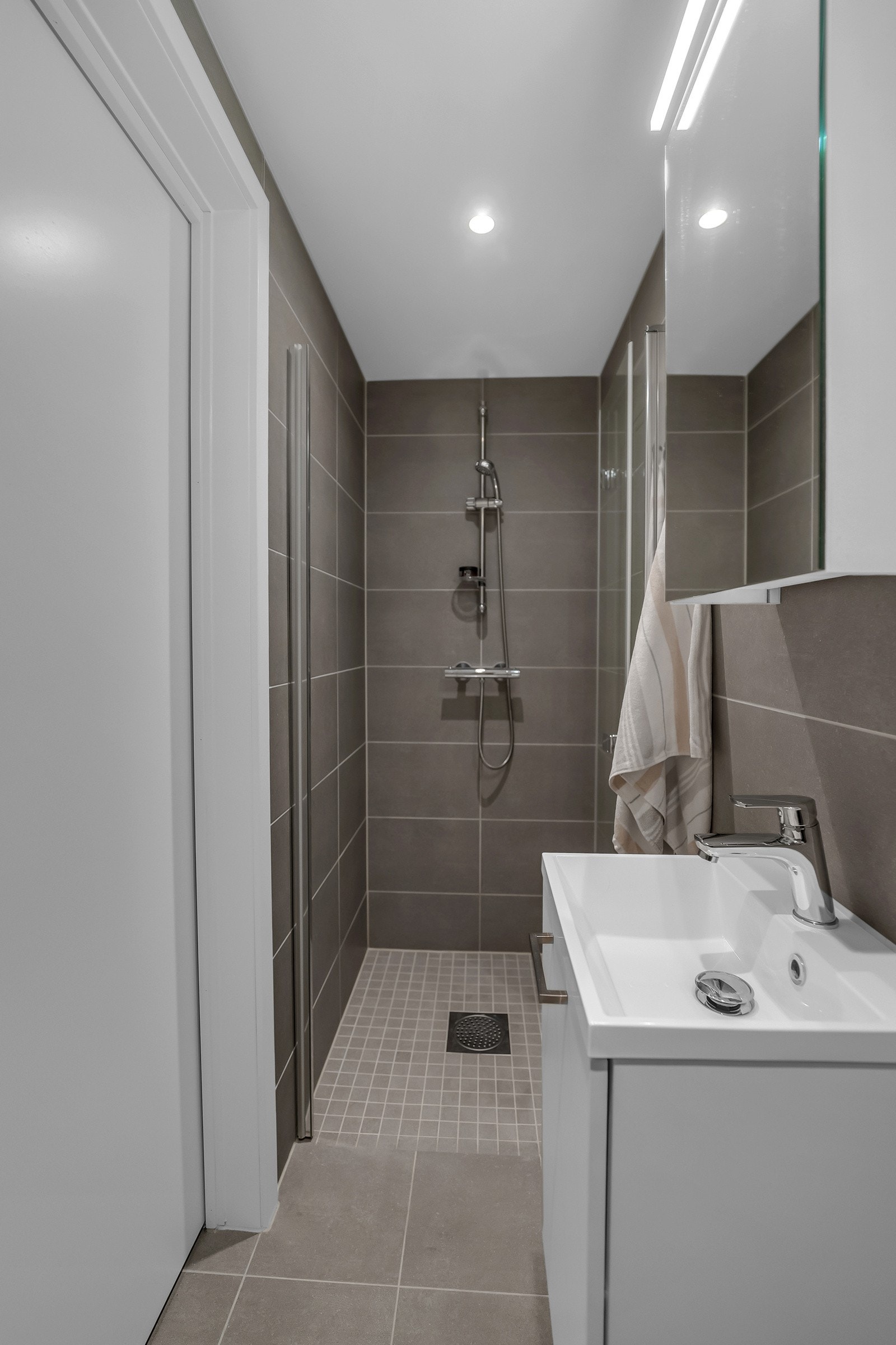 Badet har en hvit skuffeseksjon med heldekkende servant og et enkelt speilskap med god belysning. Videre er dette badet utstyrt med et vegghengt toalett og en dusjnisje med innfellbare glassdører Galleribilde