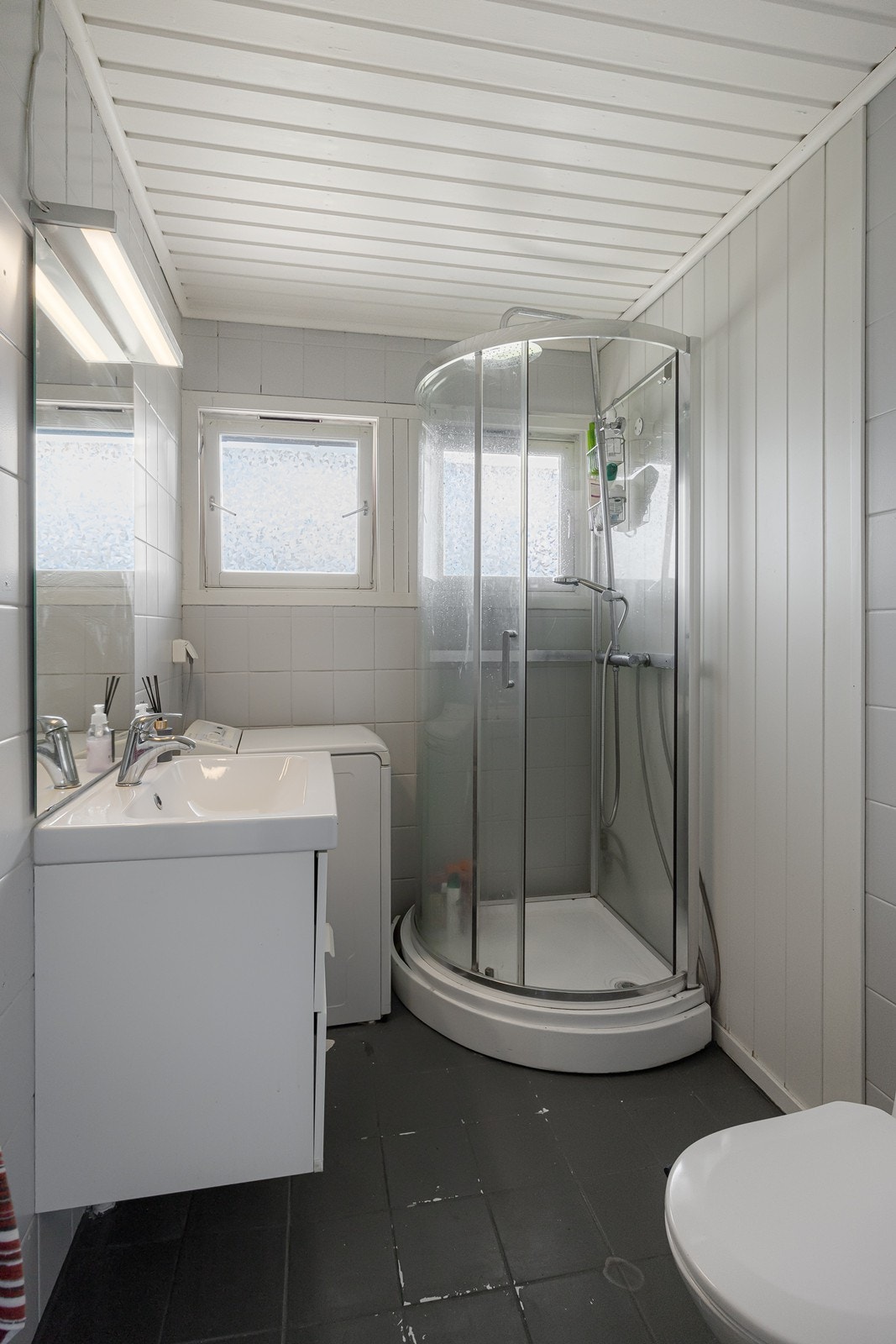 Badet har servant over skuffeseksjon og tilhørende speil med lys. Videre har badet gulvmontert toalett, dusjkabinett og opplegg for vaskemaskin. Galleribilde
