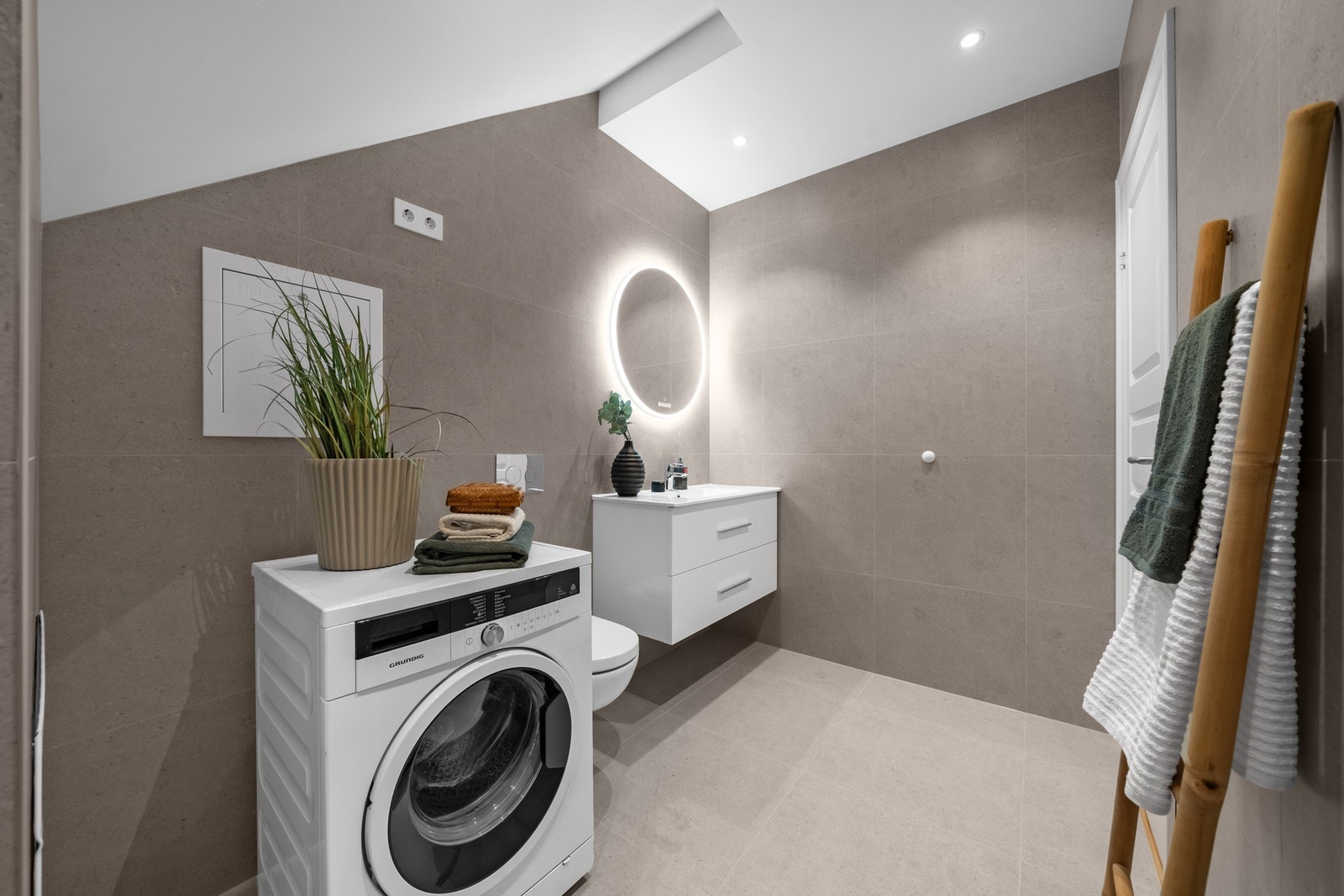 Innredning bestående av baderomsmøbel med glatte fronter, vask, moderne speil med belysning og veggfestet toalett. Det er opplegg for og plass til vaskemaskin. Rommet har avtrekk via balansert ventilasjon. Galleribilde