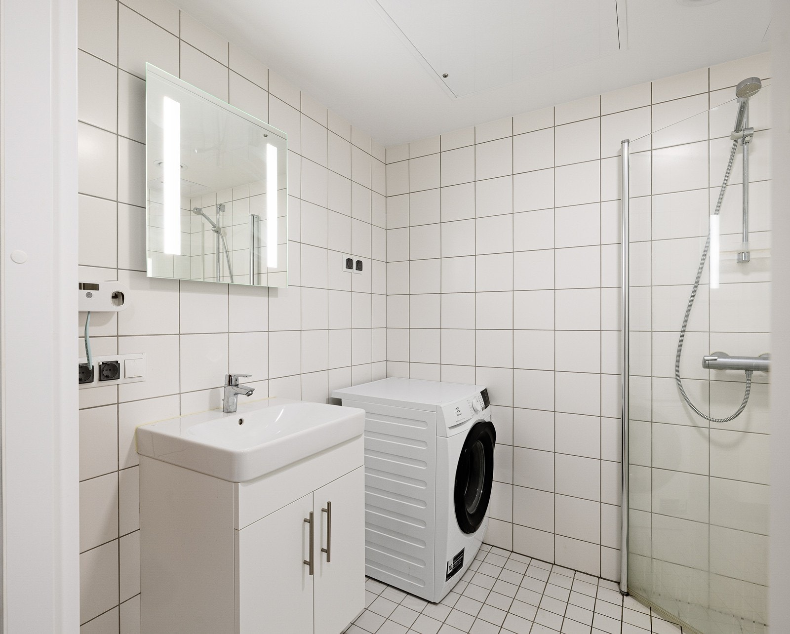 Rommet er utstyrt med vegghengt servantskap, speil med belysning og et praktisk dusjhjørne med innfellbare glassdører som sparer plass. Galleribilde