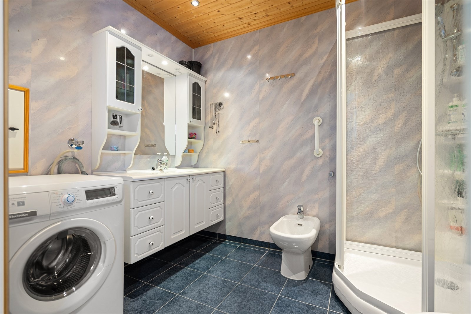 Badet inneholder opplegg for vaskemaskin, bilde, dusjkabinett samt en stor baderomsinnredning med heldekkende servant. Galleribilde