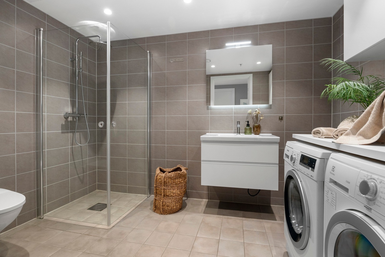 Lyst og lekkert bad med fliser og elektrisk varmekabler. Badet er utstyrt med veggmontert servant og toalett samt dushjørne med dører av glass og regnfallsdusj. Galleribilde