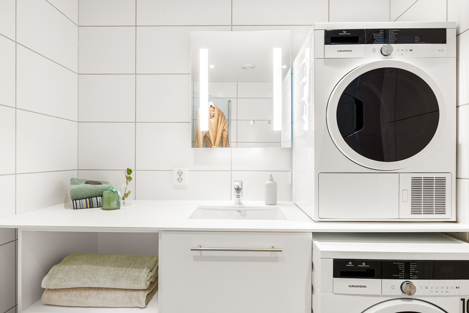 Badet inneholder innfellbare og plassbesparende dusjdører i glass, dusjbatteri og garnityr montert på vegg, kran og avløp for vaskemaskin og veggmontert WC. Rørskap er lokalisert over toalettet. Galleribilde