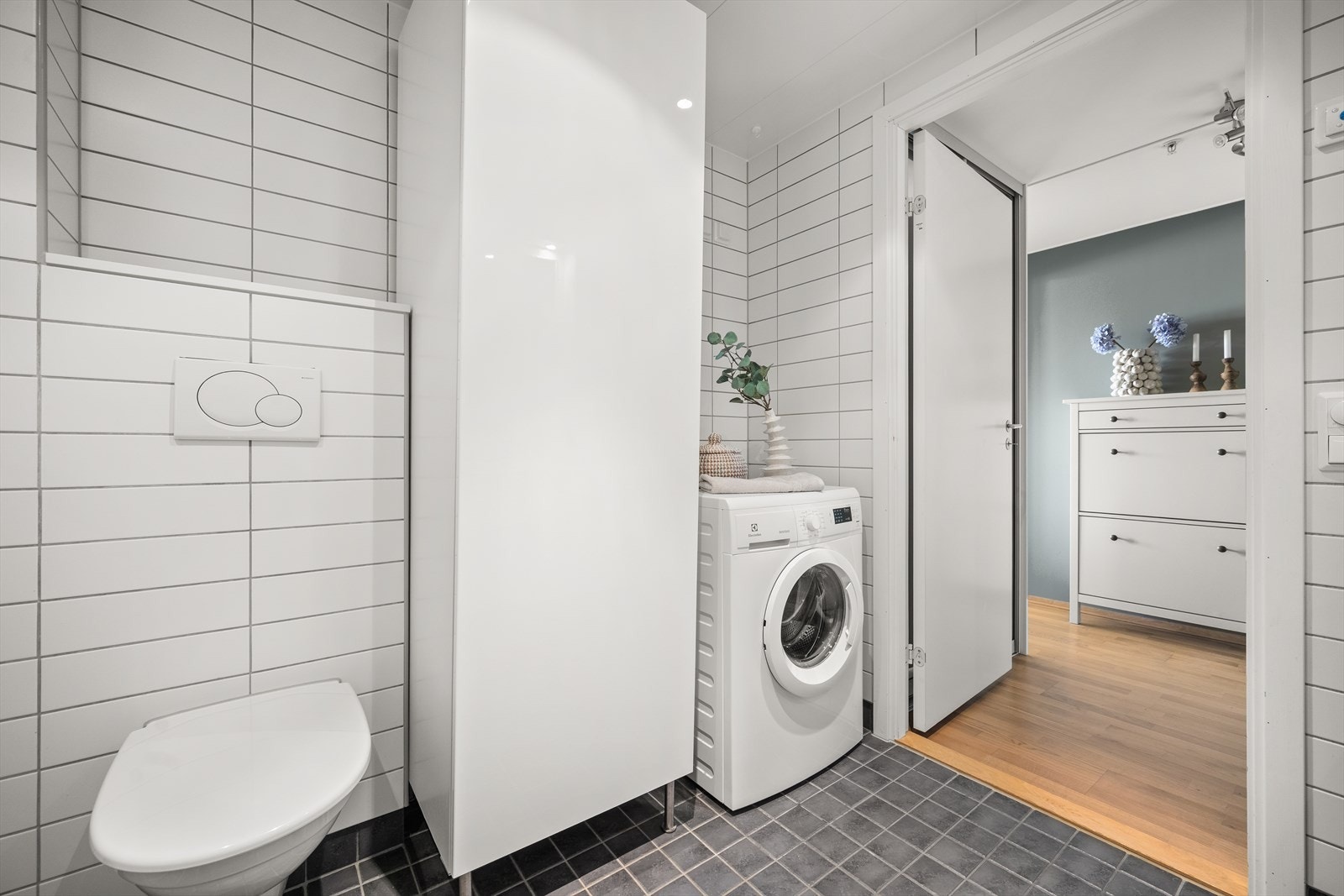 Badet er utstyrt med vegghengt toalett, dusjhjørne med innfellbare glassdører og bred baderomsinnredning med skuffer, nedfelt servant og speil med belysning. Opplegg for vaskemaskin og balansert ventilasjon. Galleribilde