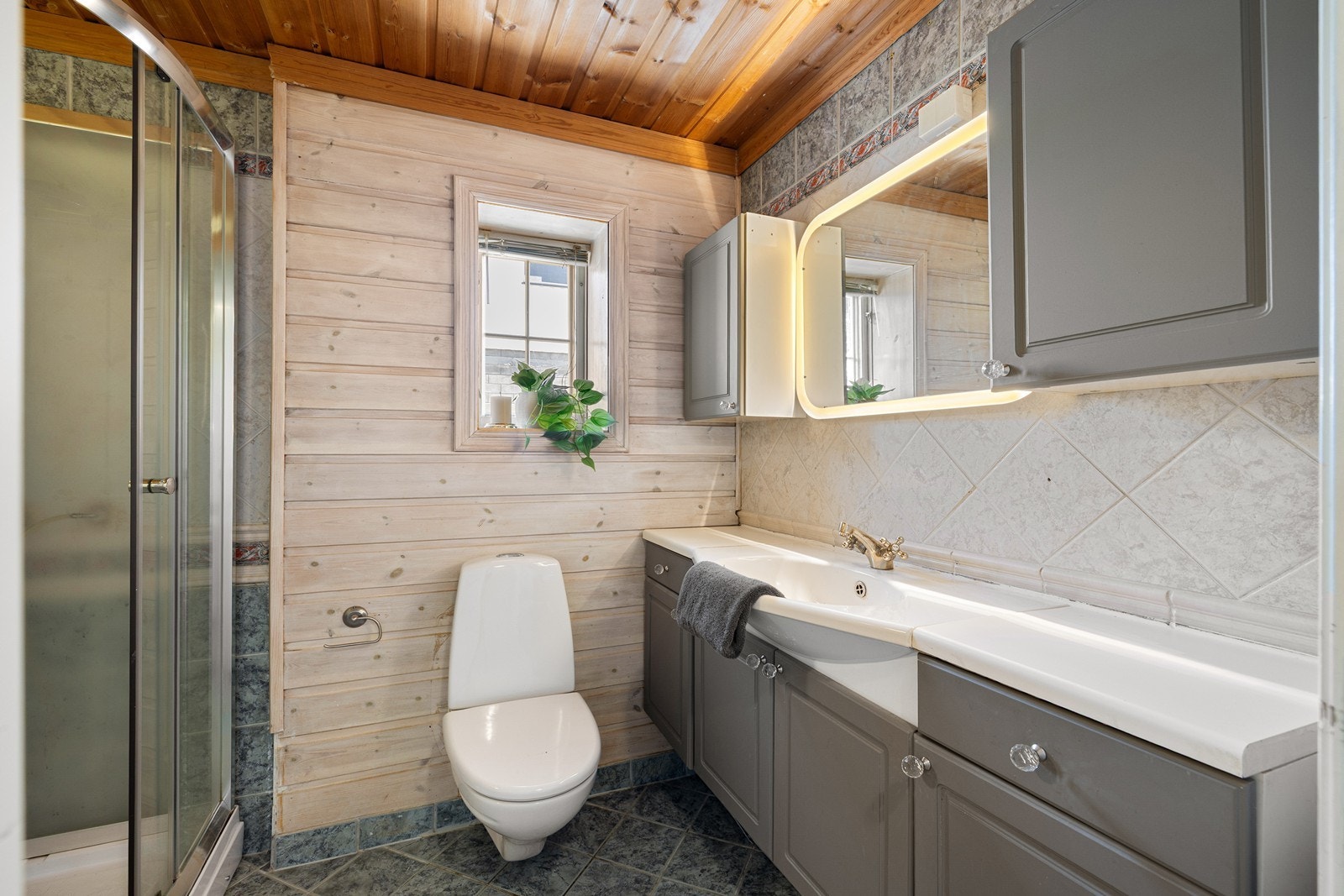 Badet i hovedetasjen har en kombinasjon av flislagte overflater og panel. Innredningen består av en moderne servant med skapseksjon og speil med integrert belysning. Galleribilde