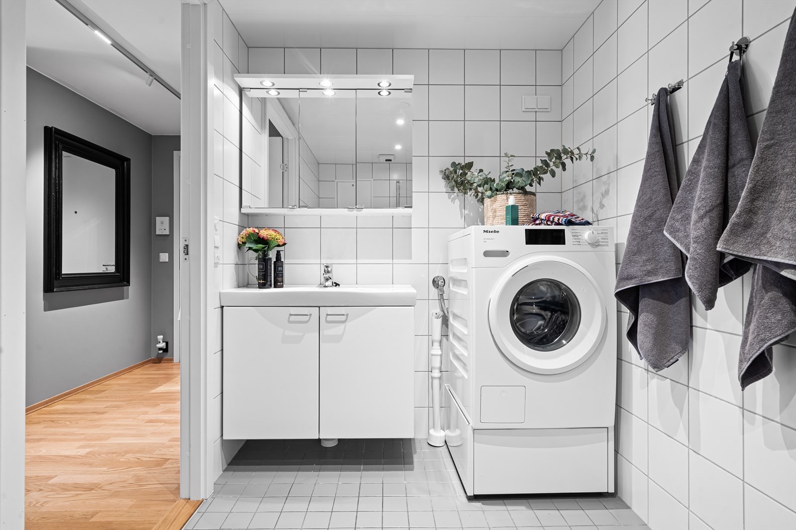 Badet er innredet med veggmontert toalett, dusj, speil med overliggende belysning, baderomsinnredning med malte glatte fronter og heldekkende servant. Videre er det opplegg for vaskemaskin og plass til tørketrommel. Galleribilde