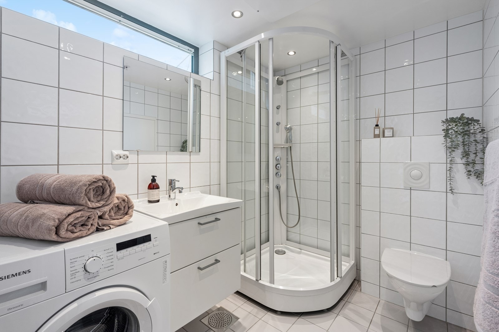 Det moderne badet er fra byggeåret og kombinerer funksjonene til bad og vaskerom. Rommet er helfliset med elektriske varmekabler. Innredningen består av servant med benkeskap, speil og et gulvmontert toalett. Dusj med glassdører, opplegg for vaskemaskin. Galleribilde