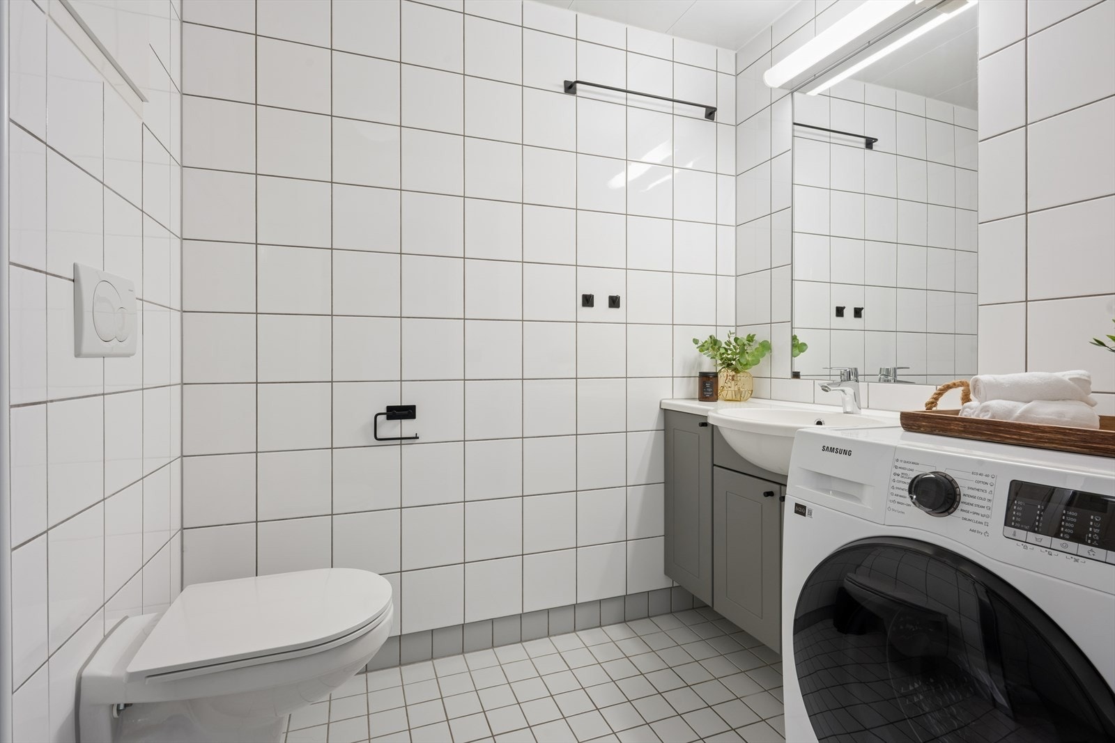 Flislagt baderom med moderne uttrykk og praktiske løsninger. Baderommet er utstyrt med vegghengt toalett, servant med underskap, dusjhjørne og opplegg til vaskemaskin. Galleribilde