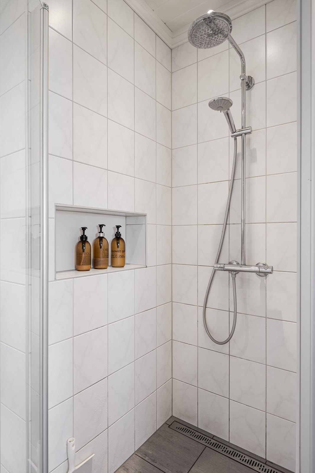 Romslig dusjnisje med glassdør, designsluk, vegghengt armatur med både hånd- og regnfallsdusj, samt innebygd hyller for såper. Galleribilde