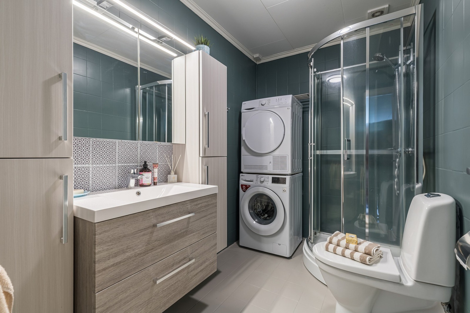 Badet har flislagte gulv og vegger, og er innredet med dusjkabinett, gulvstående toalett og baderomsinnredning med skap og skuffer for praktisk oppbevaring. Galleribilde