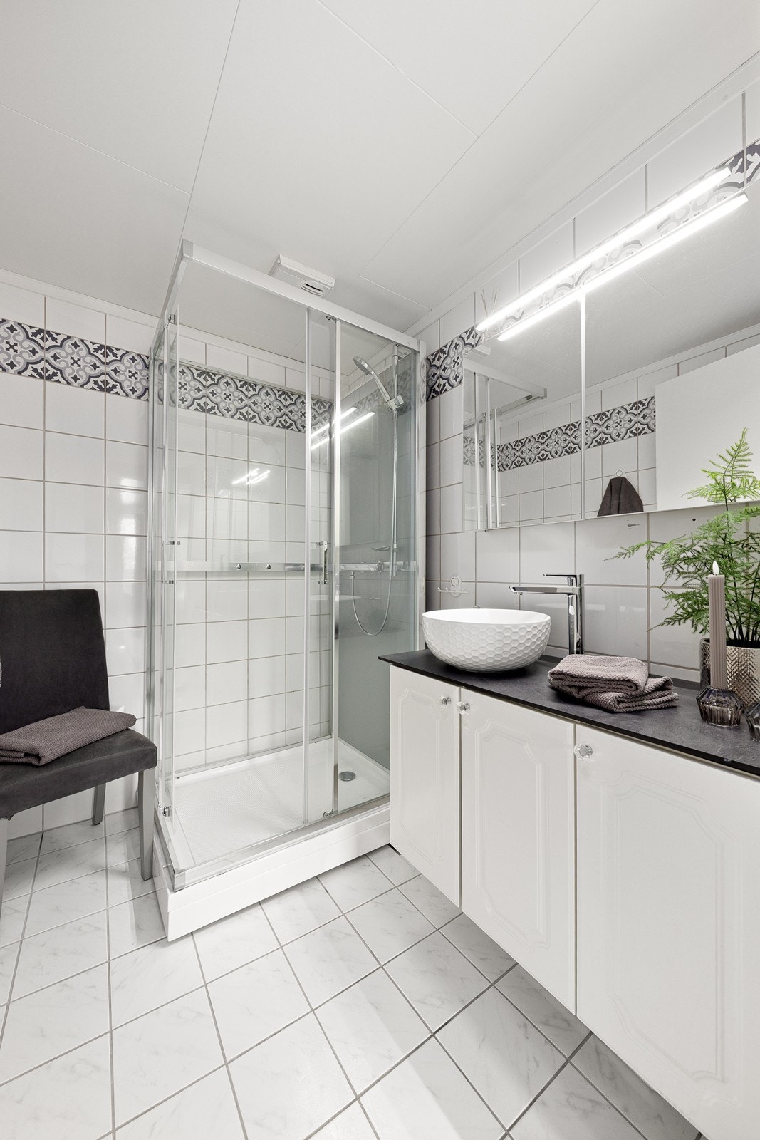 Badet har flislagt gulv med varme, dusjkabinett og en baderomsinnredning fra 2023. Rommet er av eldre dato og har et oppgraderingsbehov. Galleribilde