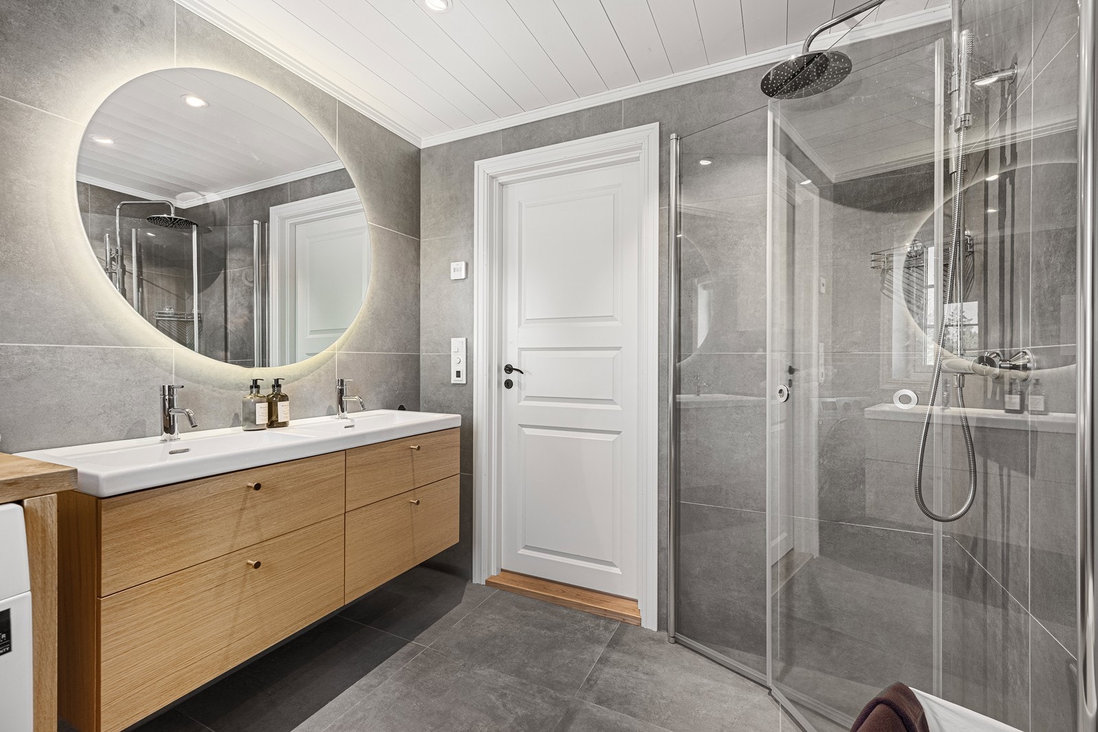 Badet ved entré er flislagt med varmekabler i gulv. Rommet inneholder dobbel servant med skuffeinnredning, samt skap i samme stil som innredningen. Videre er det opplegg for vaskemaskin, vegghengt toalett og dusjhjørne med innfellbare dusjvegger. Galleribilde