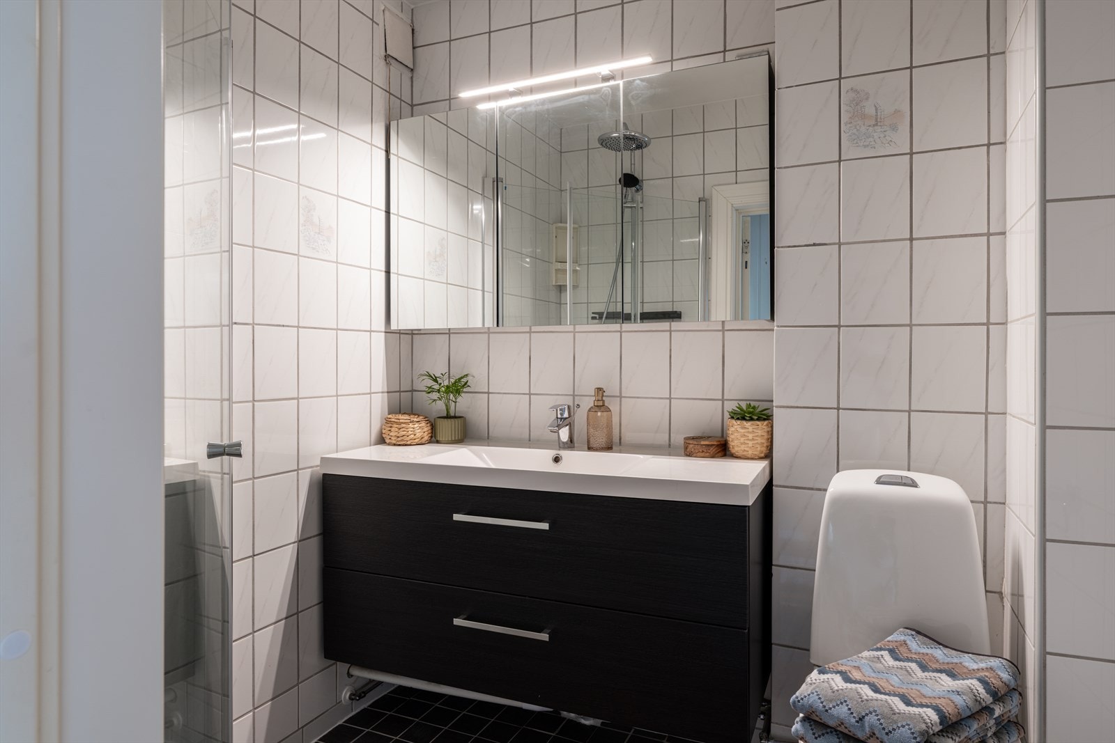 Badet er utstyrt med dusjhjørne med innsvingbare glassdører, servant med speilskap og toalett. Galleribilde