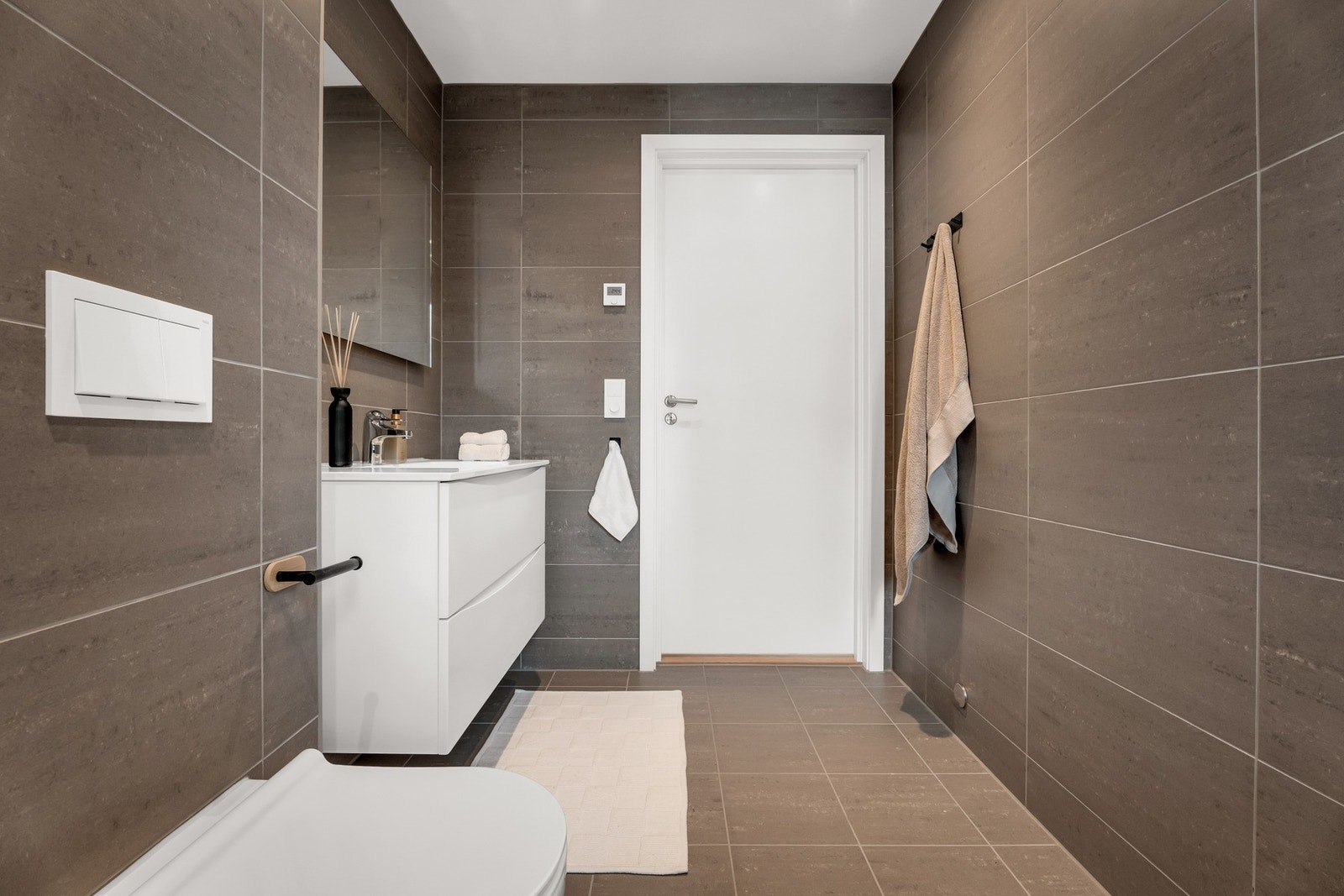 Badet inneholder flislagt dusj med glassdører, vegghengt toalett, praktisk baderomsinnredning og opplegg for vaskemaskin. Galleribilde