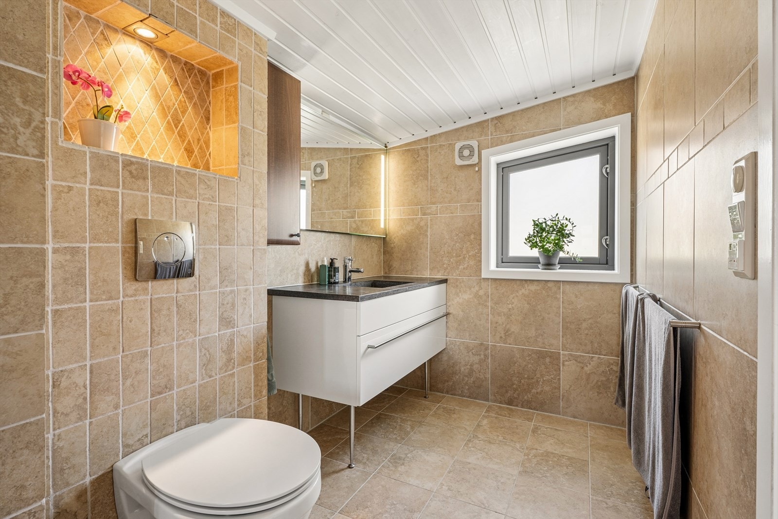 Badet i loftsetasjen er fra 2007, med fliser på gulv og vegger, gulvvarme og en praktisk, innfelt hylle. Galleribilde
