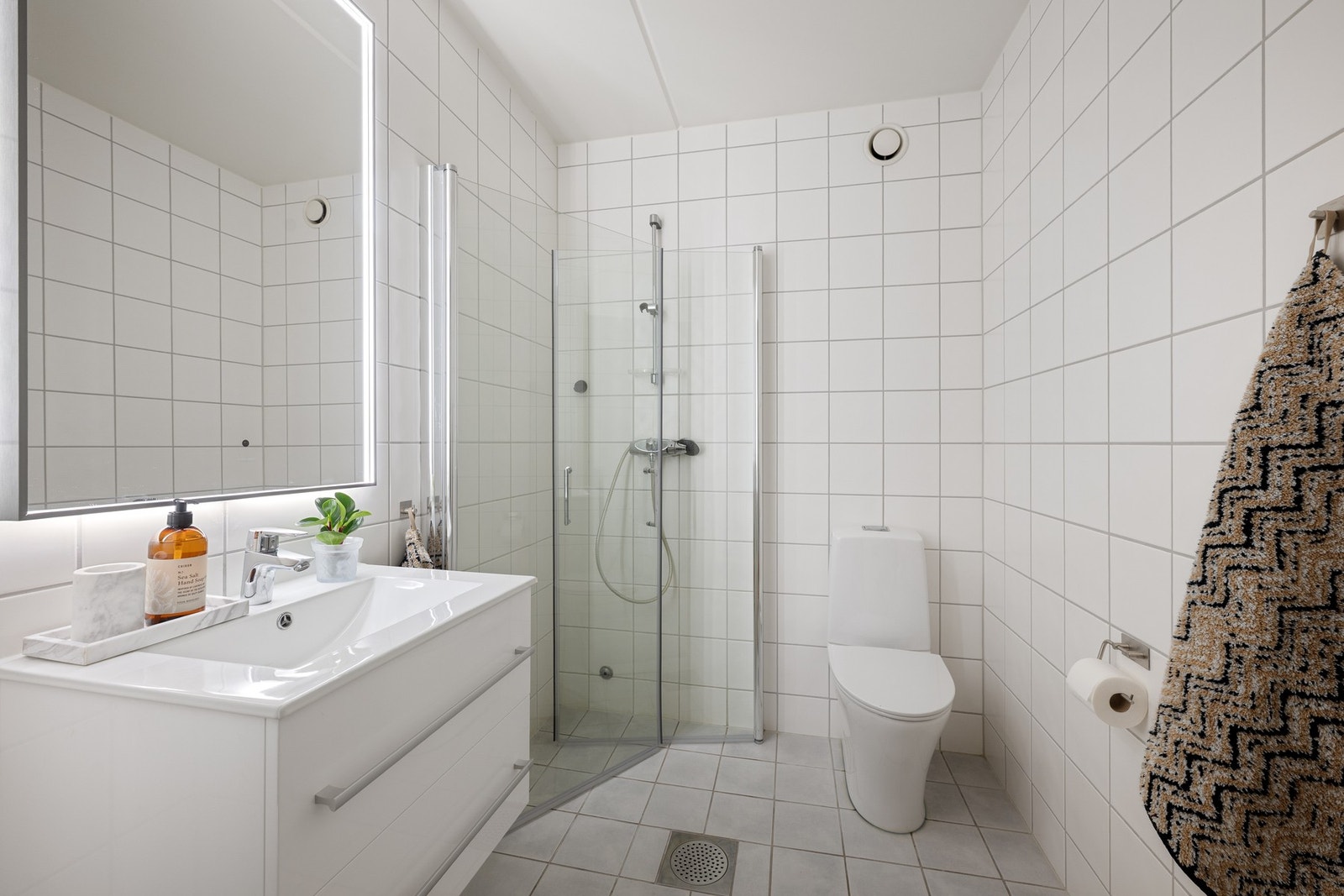 Badet med adkomst fra entré/gang er flislagt og har en praktisk utforming med dusjhjørne, gulvmontert toalett og servantinnredning med speilbelysning. Rommet har varmekabler i gulv og mekanisk avtrekk. Galleribilde