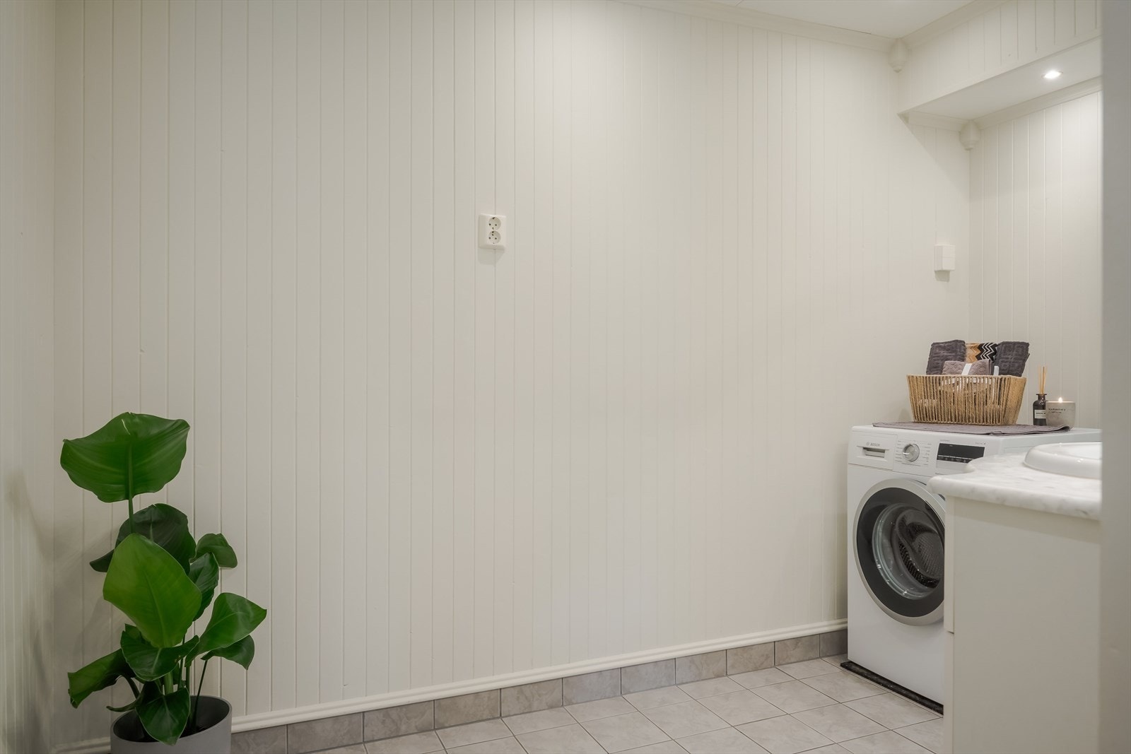 Lyst bad/vaskerom med overflater, installasjoner og komponenter fra perioden ca. 2005 til 2007. Baderommet har flislagt gulv med gulvvarme, panel på veggene som ble malt med våtromsmaling i 2025, downlights i himlingen og elektrisk styrt vifte. Galleribilde