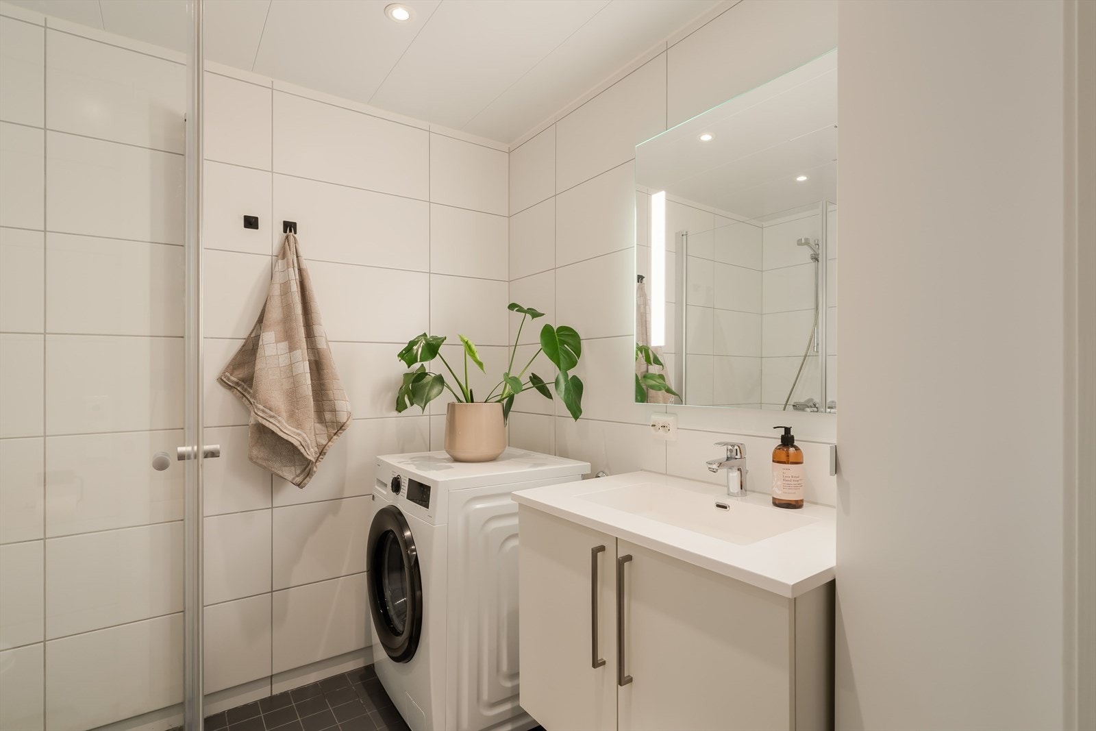 Det inneholder dusjvegger av glass, vegghengt toalett, innredning fra HTH med servant i benkeskap, og opplegg for vaskemaskin. Galleribilde