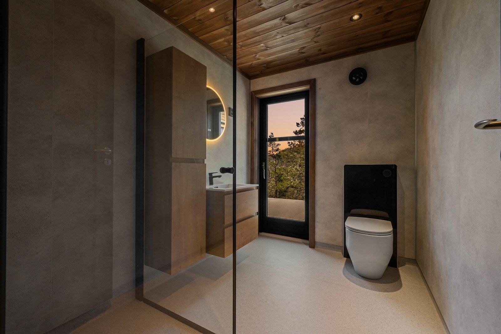 Badet er stilrent med moderne overflater og varme detaljer i tre. Badet er innredet med servantskap med slette trefargede fronter, heldekkende servant og sort armatur. I tillegg er det montert høyskap som gir praktisk oppbevaringsplass, samt rundt speil m Galleribilde