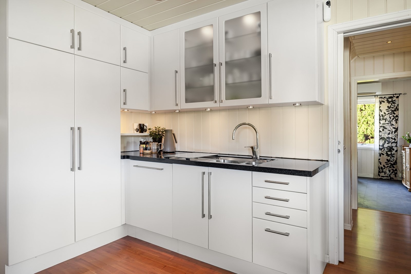 Kjøkkenet har hvite, glatte fronter og enkelte overskap med fronter med glass. Benkeplaten er av stein med nedfelt oppvaskkum i rustfritt stål med liten skyllekum. Galleribilde
