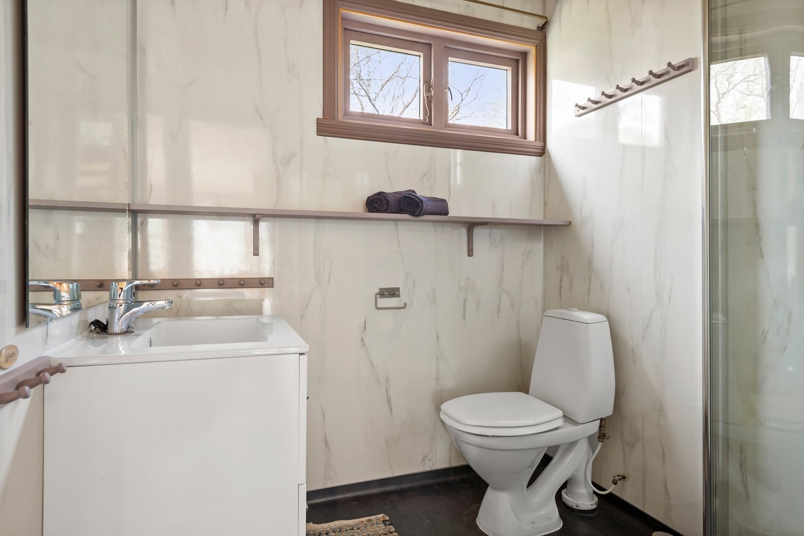 Badet har vinylbelegg på gulv med elektriske varmekabler, baderomsplater på veggene og panel i taket. Rommet er utstyrt med en innredning med slette fronter og nedfelt servant, toalett og dusjhjørne Galleribilde