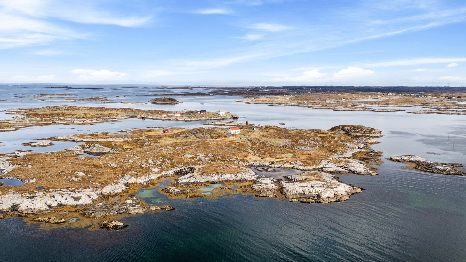 Dronebilde over holmen og nærliggende omgivelser, sett nordover. Mot nordøst ser man vindturbinparken på Smøla. Galleribilde