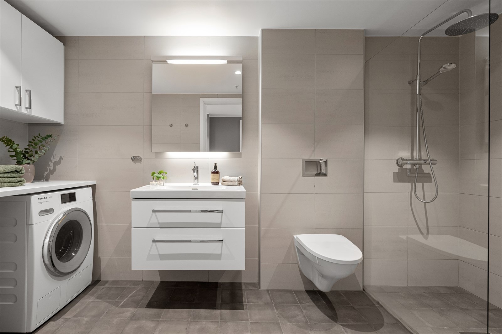 Badet er svært delikat - flislagt med fliser i duse toner på gulv og vegg. Badet er innredet med vegghengt toalett, servant i skuffeinnredning, dusj med fast dusjvegg , opplegg for vaskemaskin og tørketrommel i tillegg til en praktisk vaskeromsinnredning. Galleribilde