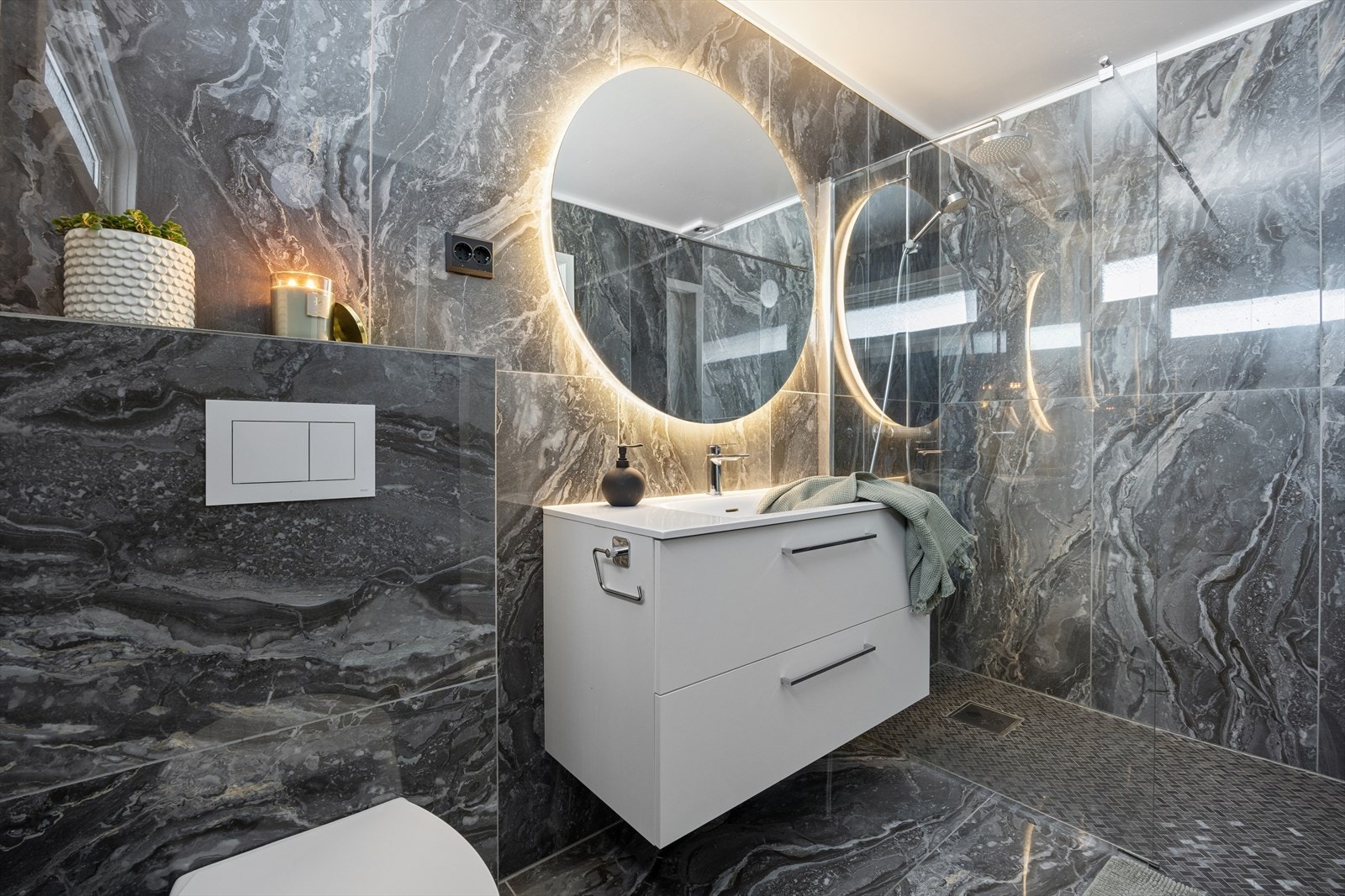 Badet er oppgradert i 2023 og er delikat innredet med store, mørke fliser på gulv og vegger. I gulvet har man også varmekabler for ekstra komfort. Badet er ellers utstyrt med dusjhjørne, vegghengt toalett og baderomsinnredning med nedfelt servant. Galleribilde